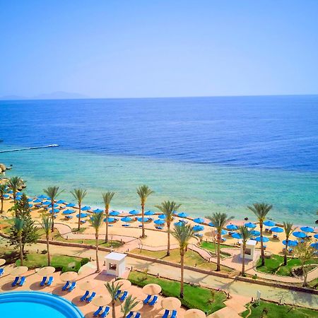 فندق رويال جراند شرم ريزورت شرم الشيخ المظهر الخارجي الصورة The photo displays a beautiful coastal scene featuring a clear blue sea that transitions into green tones near the shore. In the foreground, there are sandy beaches lined with palm trees and a variety of beach umbrellas. There is also a swimming pool