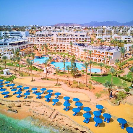 فندق رويال جراند شرم ريزورت شرم الشيخ المظهر الخارجي الصورة The photo shows a vibrant resort area with several buildings that appear to be accommodations or hotel units. There are multiple swimming pools surrounded by palm trees, creating a tropical atmosphere. In the foreground, there is a beach area with sa