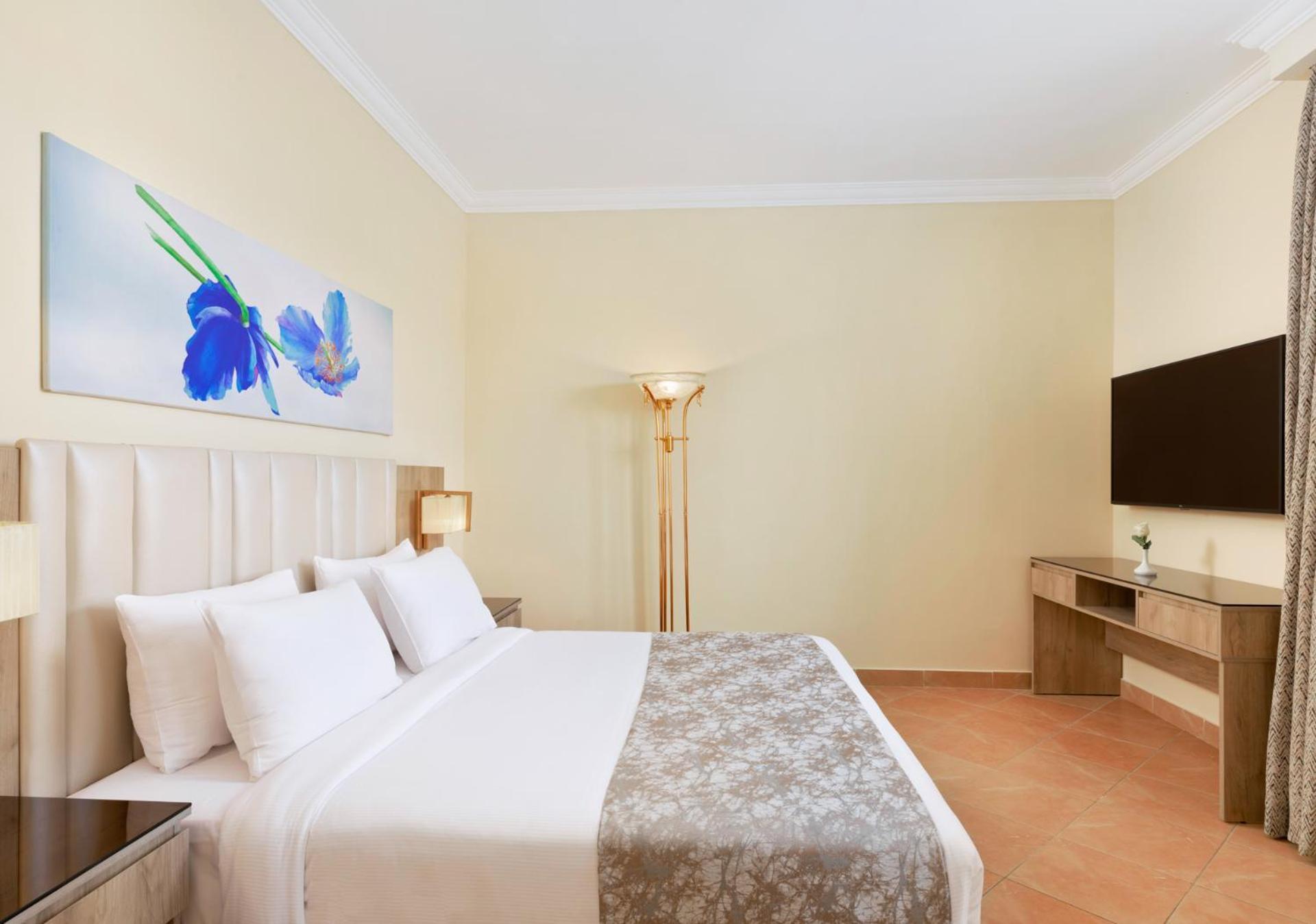 فندق رويال جراند شرم ريزورت شرم الشيخ المظهر الخارجي الصورة The photo shows a modern hotel room featuring a large bed with white linens and a decorative gray throw. On one side of the room, there is a piece of artwork depicting blue flowers. A floor lamp provides additional lighting, and across from the bed i