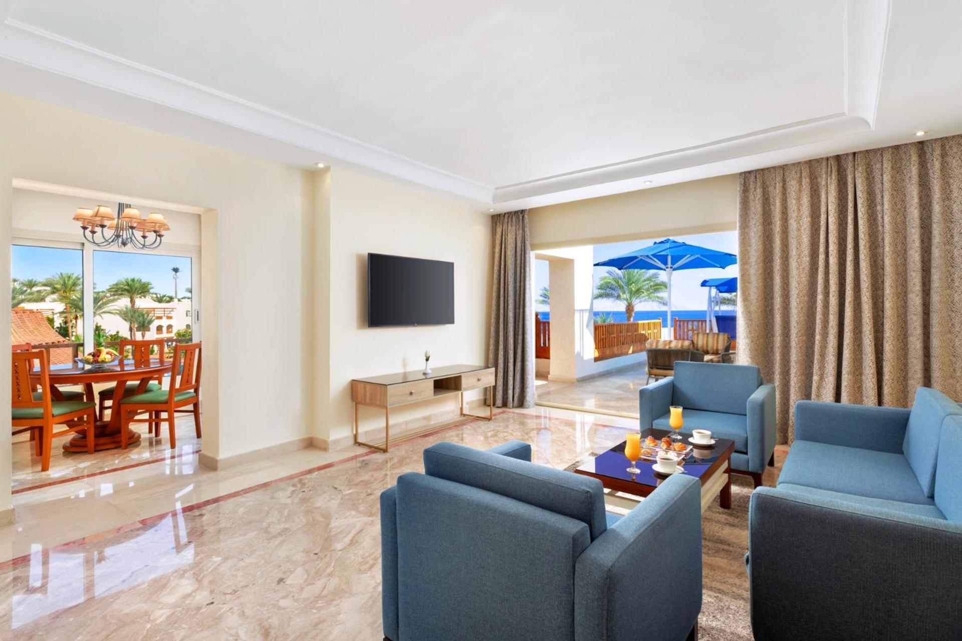فندق رويال جراند شرم ريزورت شرم الشيخ المظهر الخارجي الصورة The photo shows a modern and spacious living area. There are several blue chairs arranged around a coffee table, which holds some refreshments. A large flat-screen TV is mounted on the wall, and there are sliding glass doors that open to an outdoor a