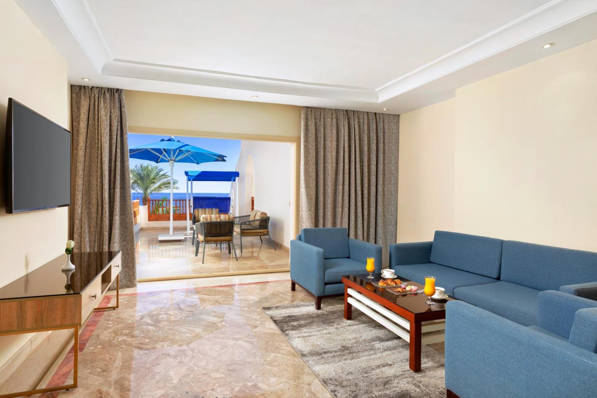 فندق رويال جراند شرم ريزورت شرم الشيخ المظهر الخارجي الصورة The photo shows a spacious living area with modern decor. There are two blue sofas arranged around a coffee table that holds some decorative items and candles. A flat-screen TV is mounted on the wall across from the seating area. To the left, there a
