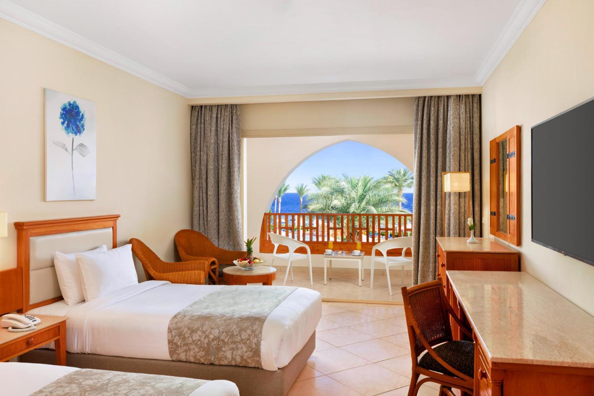 فندق رويال جراند شرم ريزورت شرم الشيخ المظهر الخارجي الصورة The photo shows a hotel room featuring two beds with light bedding. There is a wooden desk and chair, and a comfortable chair is placed nearby. The room has large windows that open to a balcony with a view of palm trees and a blue sky. The decor is r
