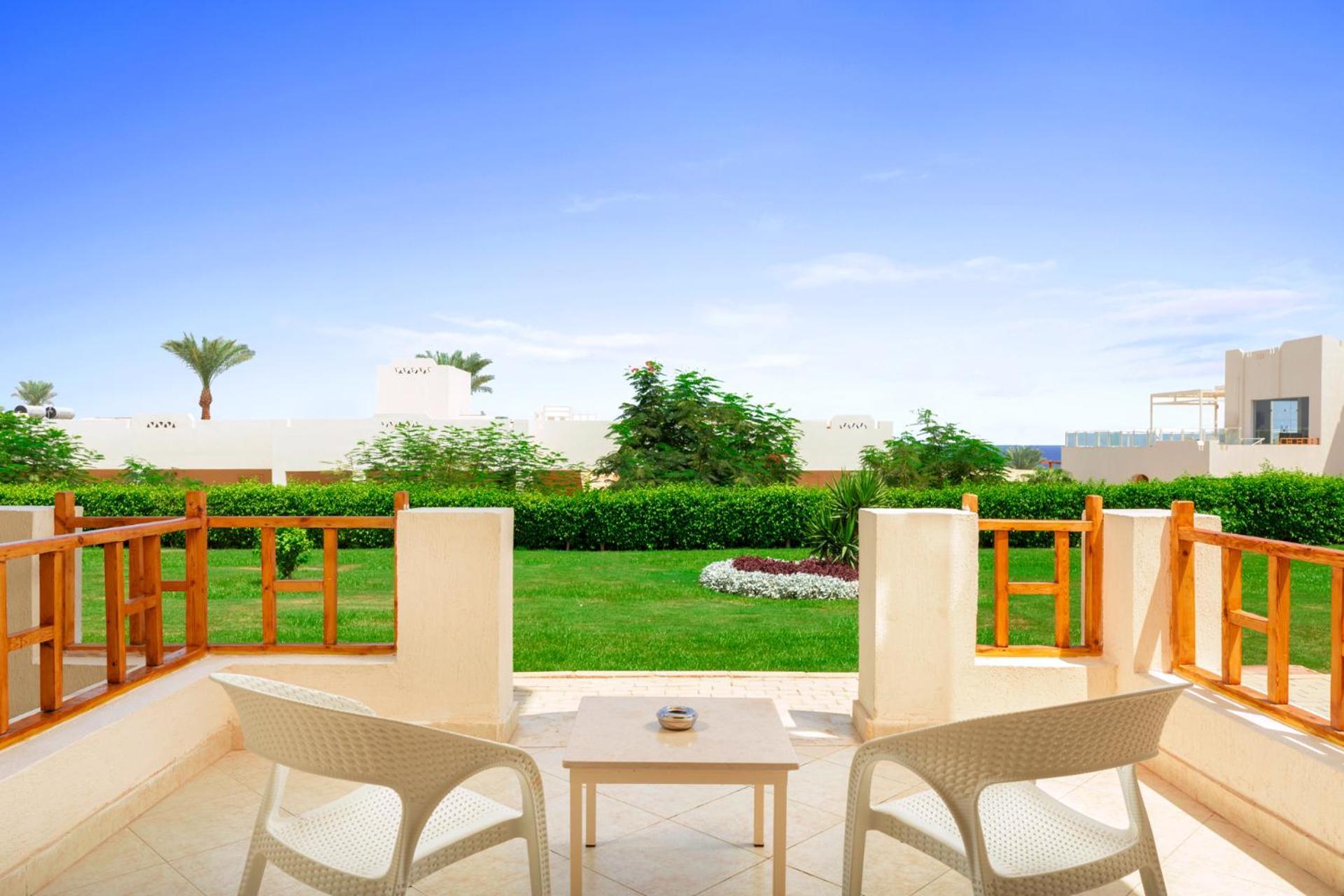 فندق رويال جراند شرم ريزورت شرم الشيخ المظهر الخارجي الصورة The photo depicts a serene outdoor balcony or patio area. It features two white chairs and a small table, set against a backdrop of lush green grass and garden plants. In the distance, there are some palm trees and clear blue skies, suggesting a warm