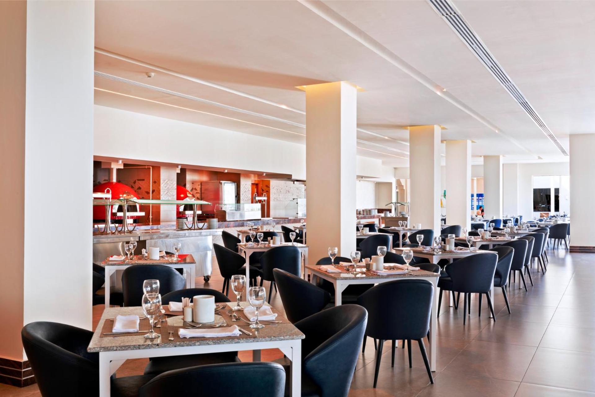 فندق رويال جراند شرم ريزورت شرم الشيخ المظهر الخارجي الصورة The photo shows a modern restaurant interior. It features a spacious layout with several dining tables arranged in a row, each set with dishes, glasses, and cutlery. The tables have light-colored surfaces and are accompanied by black chairs. In the b