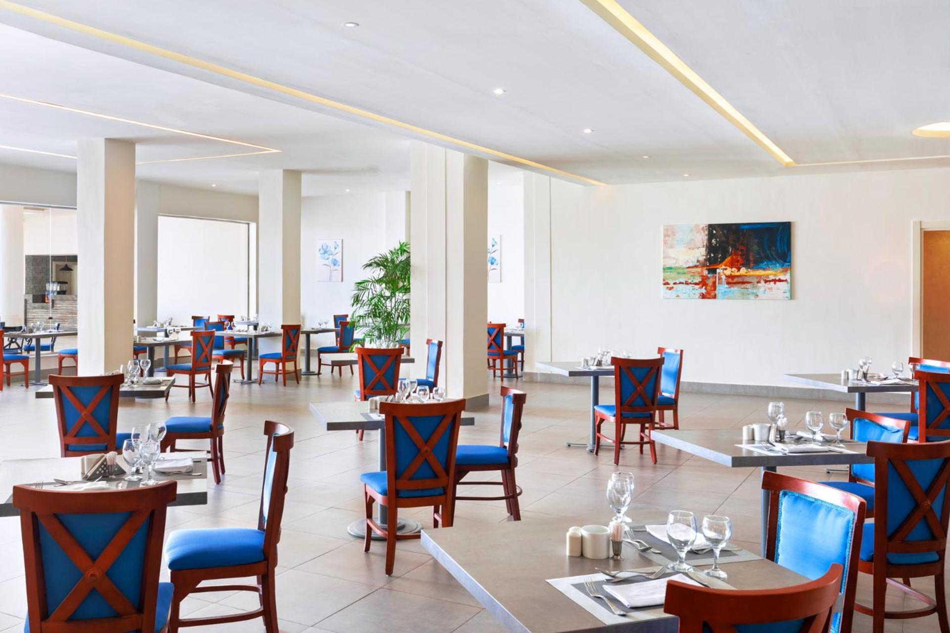 فندق رويال جراند شرم ريزورت شرم الشيخ المظهر الخارجي الصورة The photo shows an interior of a restaurant featuring a modern and spacious design. The floors are tiled, and the walls are painted in neutral tones. The seating consists of wooden chairs with blue cushions arranged around tables, which are set with 