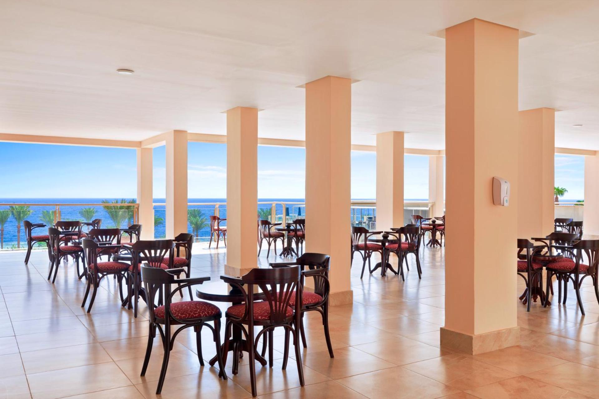 فندق رويال جراند شرم ريزورت شرم الشيخ المظهر الخارجي الصورة The image shows a dining area or café with several round wooden tables and chairs, featuring red upholstery. The space is well-lit, with large windows that allow a view of the ocean in the background, contributing to a bright and open atmosphere. The