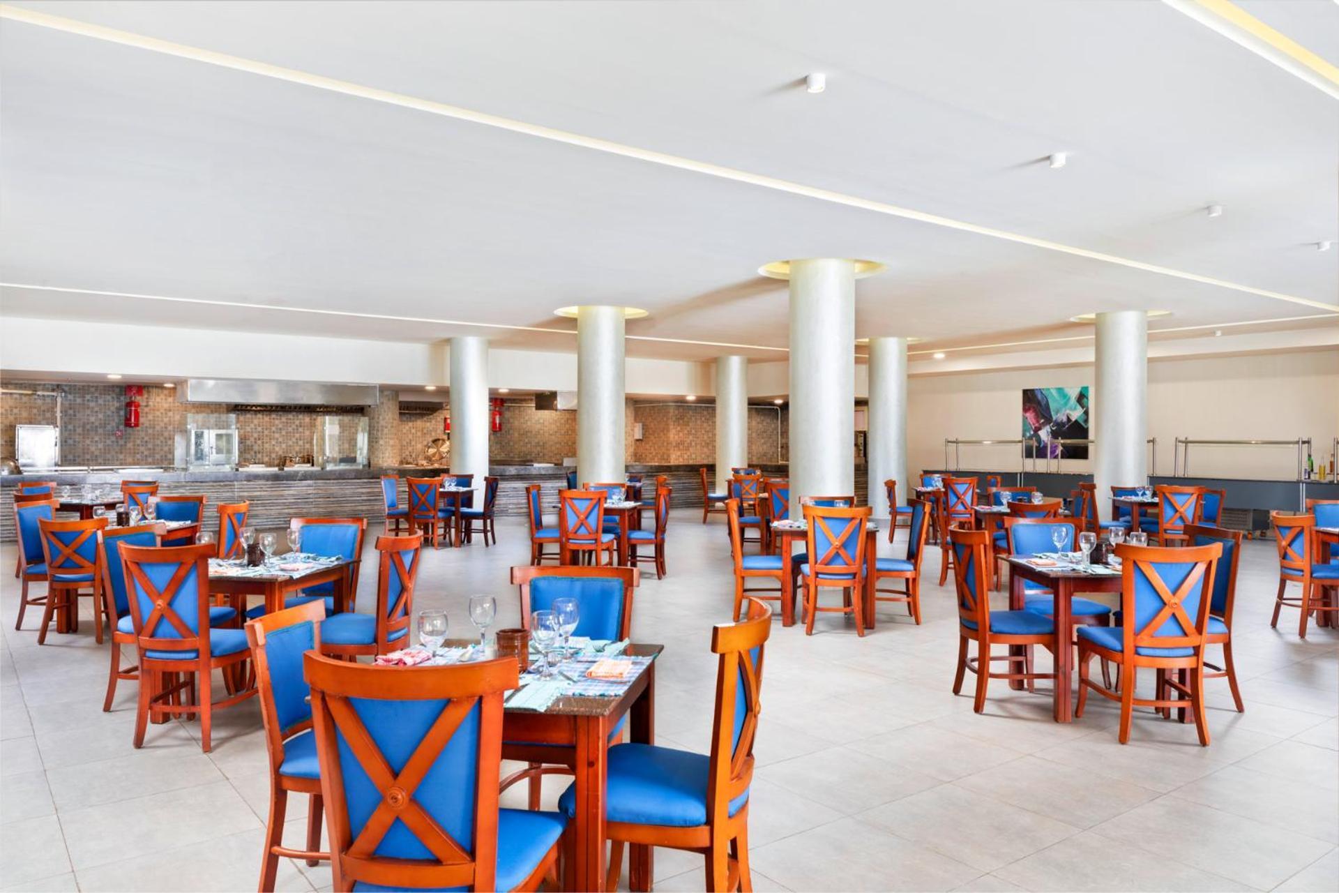 فندق رويال جراند شرم ريزورت شرم الشيخ المظهر الخارجي الصورة The photo depicts a spacious dining area in a restaurant. The room features several wooden tables, each with blue chairs. The chairs have a unique design with prominent X-shaped backs. The flooring is light-colored tile, and the walls are painted in 