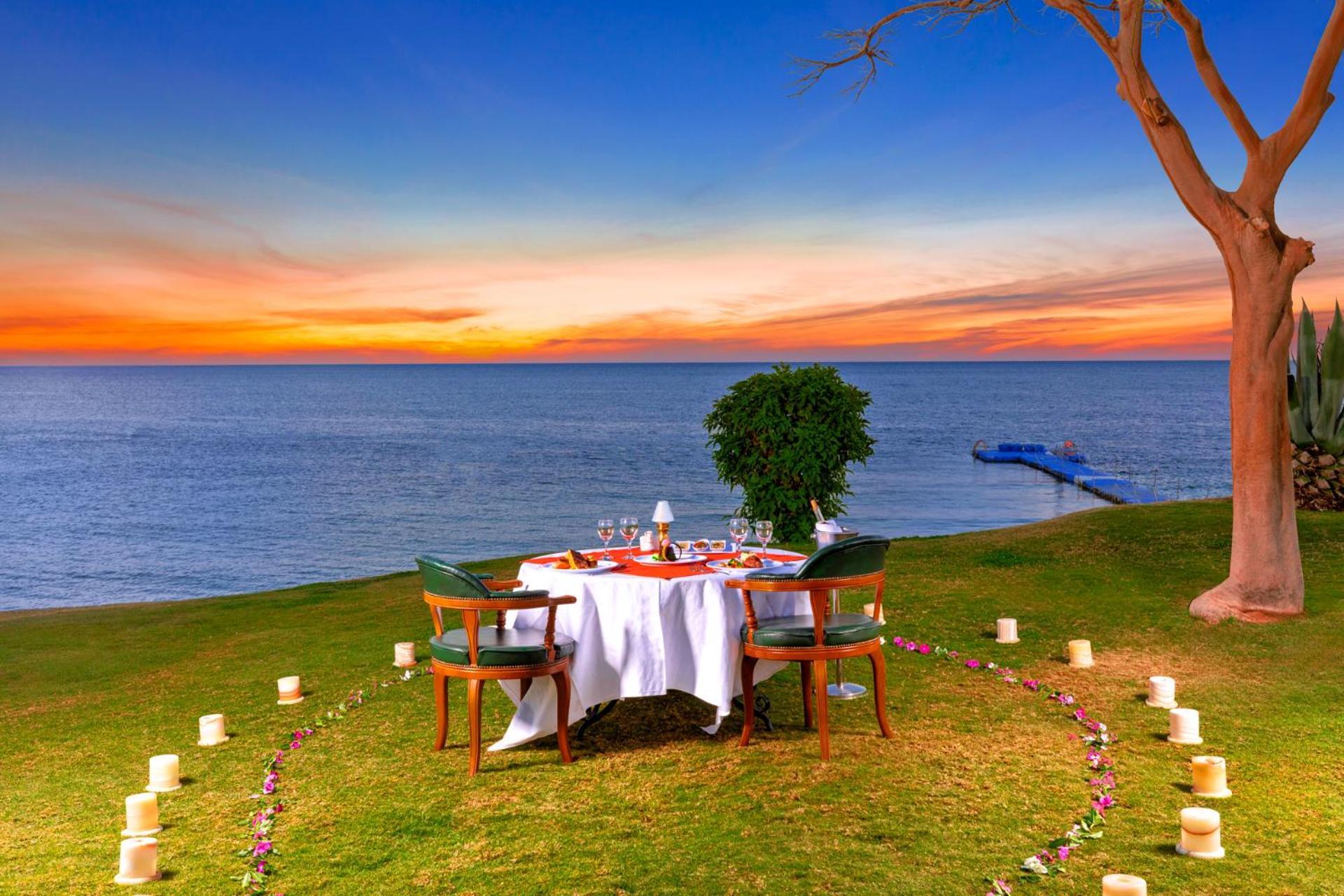 فندق رويال جراند شرم ريزورت شرم الشيخ المظهر الخارجي الصورة The photo depicts a romantic outdoor dining setup by the seaside. In the foreground, there is a round table elegantly set with a white tablecloth, plates, and glassware, creating a cozy atmosphere. Surrounding the table are green chairs. A circle of 
