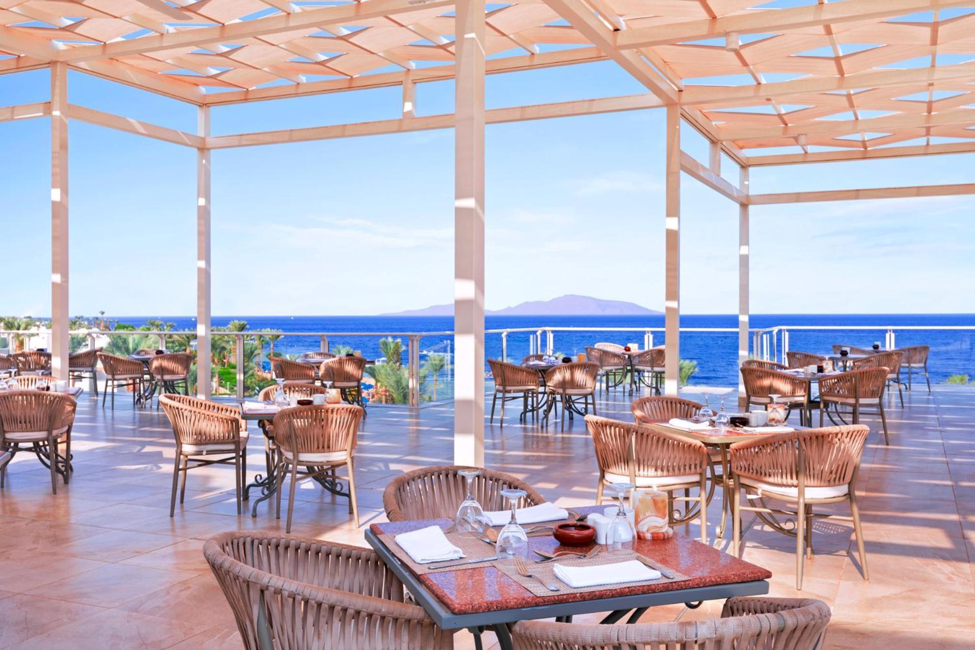 فندق رويال جراند شرم ريزورت شرم الشيخ المظهر الخارجي الصورة The photo shows a spacious outdoor restaurant with a beautiful view of the ocean. The seating area features several tables and wicker chairs. The open design includes a wooden pergola providing some shade, while large windows or openings allow for a 
