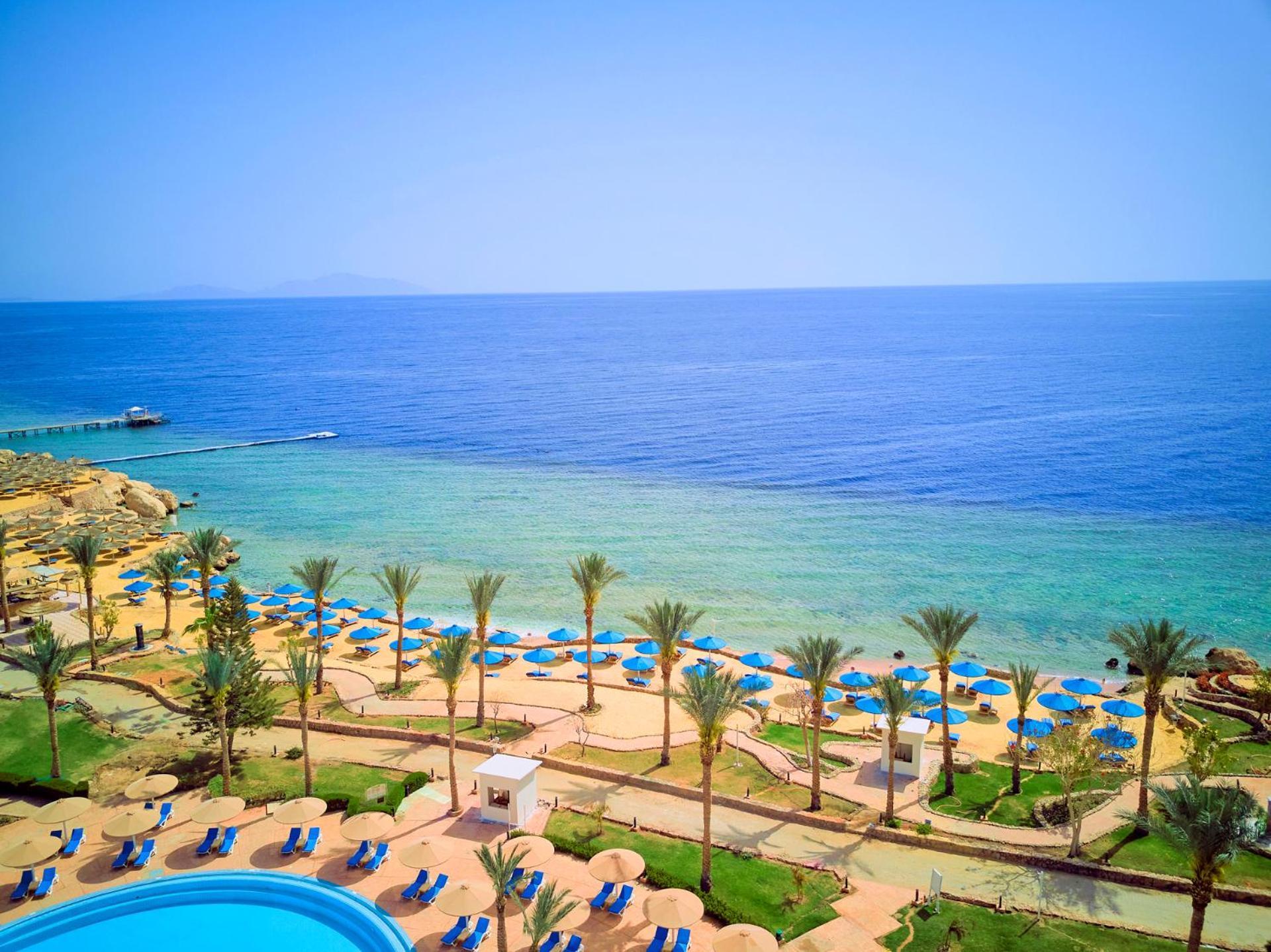 فندق رويال جراند شرم ريزورت شرم الشيخ المظهر الخارجي الصورة The photo displays a beautiful coastal scene featuring a clear blue sea that transitions into green tones near the shore. In the foreground, there are sandy beaches lined with palm trees and a variety of beach umbrellas. There is also a swimming pool