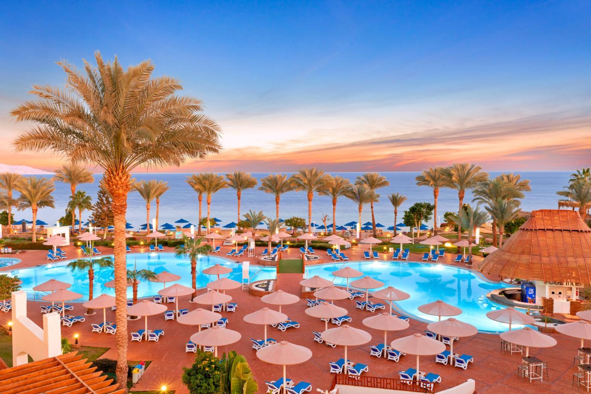 فندق رويال جراند شرم ريزورت شرم الشيخ المظهر الخارجي الصورة The image shows a beautiful resort setting by the beach. There is a large swimming pool at the center, surrounded by lounge chairs and umbrellas. Palm trees line the area, giving it a tropical vibe. In the background, you can see the ocean and a colo