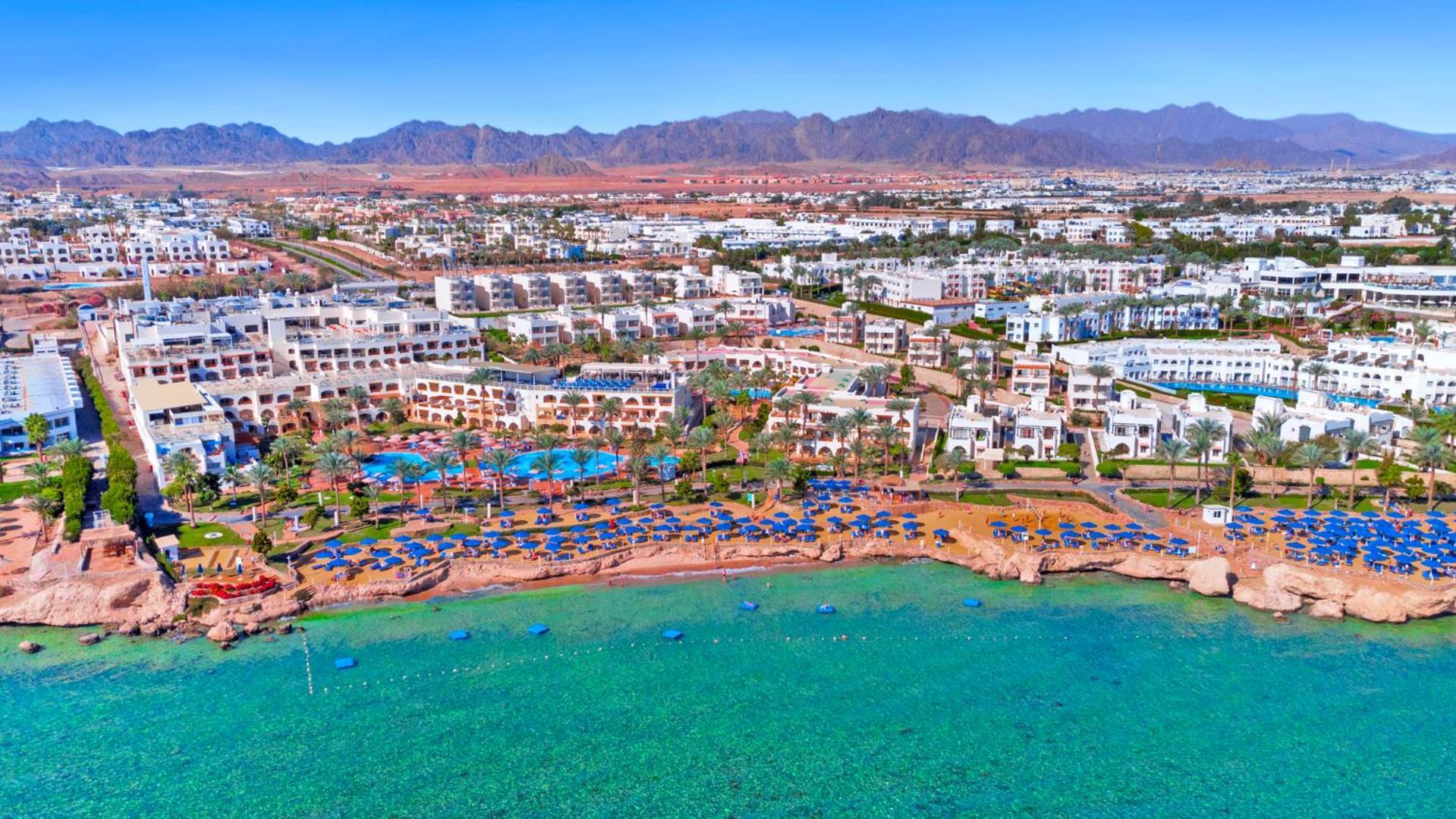 فندق رويال جراند شرم ريزورت شرم الشيخ المظهر الخارجي الصورة The photo shows an aerial view of a resort located by the coast. There are several swimming pools surrounded by sun loungers and umbrellas, as well as a beach area with clear turquoise water. In the background, there are multiple buildings of the res