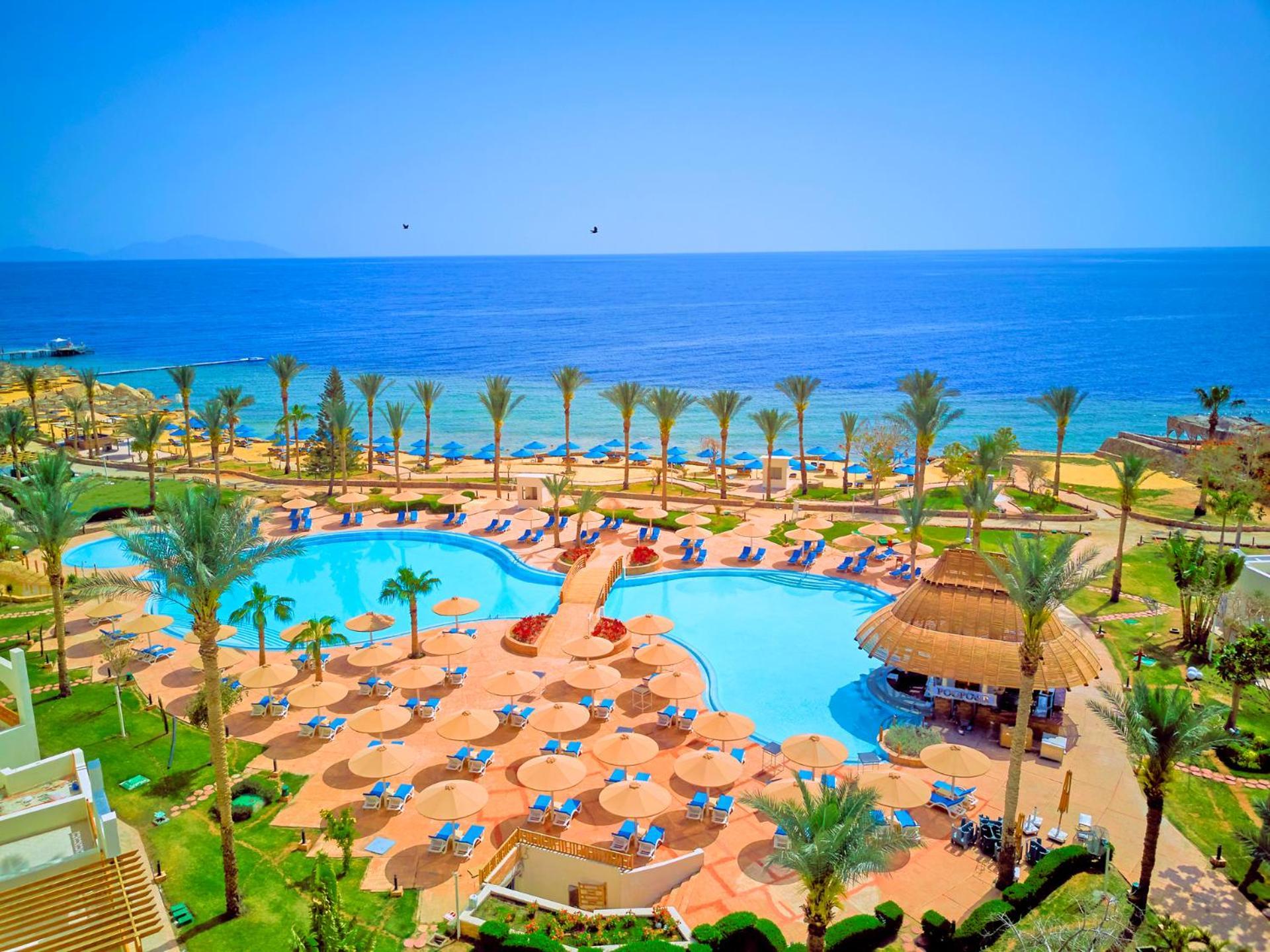 فندق رويال جراند شرم ريزورت شرم الشيخ المظهر الخارجي الصورة The photo depicts a picturesque resort scene by the sea. In the foreground, there is a large, inviting swimming pool surrounded by lounge chairs and umbrellas. Palm trees are strategically placed around the pool area, adding to the tropical atmospher