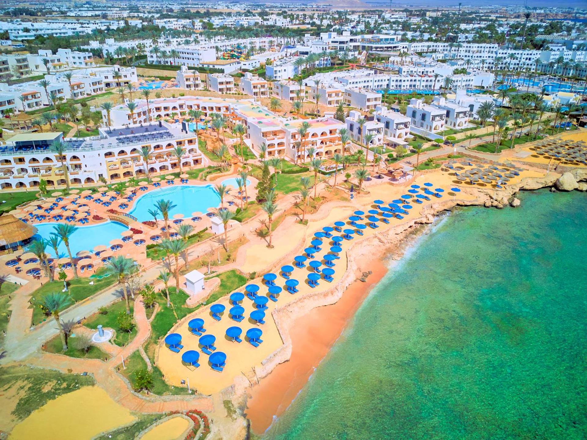 فندق رويال جراند شرم ريزورت شرم الشيخ المظهر الخارجي الصورة The image shows an aerial view of a resort area by the beach. There is a swimming pool in the center, surrounded by palm trees and sun loungers. The sandy beach features numerous umbrellas arranged in rows for shade. In the background, there are seve
