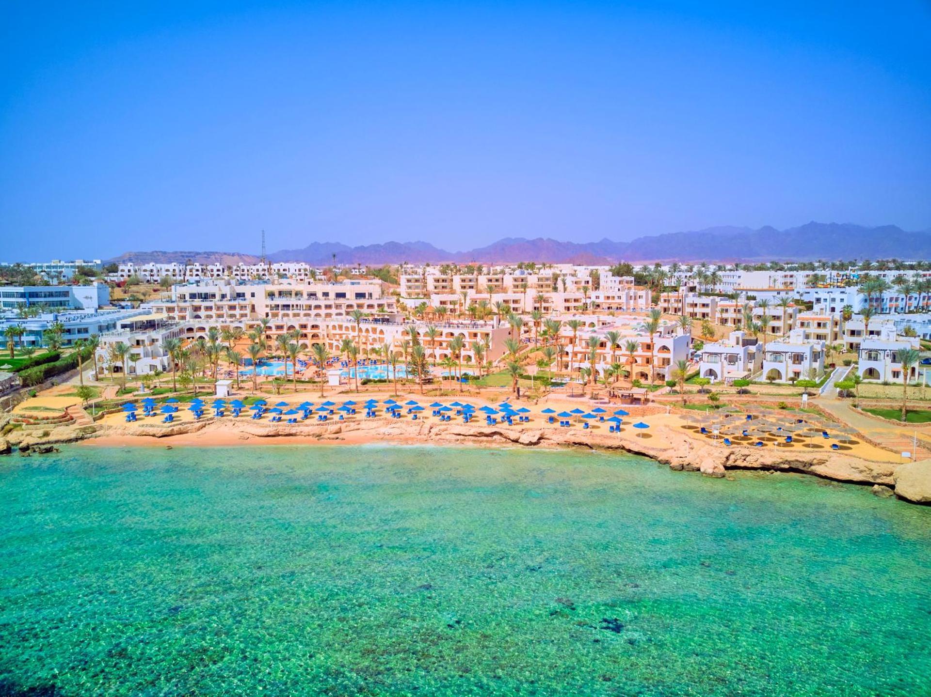 فندق رويال جراند شرم ريزورت شرم الشيخ المظهر الخارجي الصورة The photo depicts a picturesque beachfront resort. It shows a clear turquoise sea with a sandy shore lined with beach umbrellas and sun loungers. Behind the beach, there are several white buildings of the resort, surrounded by lush greenery. In the b