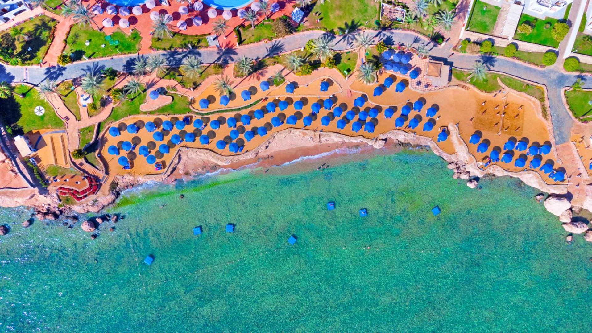 فندق رويال جراند شرم ريزورت شرم الشيخ المظهر الخارجي الصورة The photo shows an aerial view of a beach resort area. You can see a stretch of golden sandy beach lined with numerous blue umbrellas. The beach is bordered by clear turquoise waters. There are palm trees scattered throughout the area, adding to the 