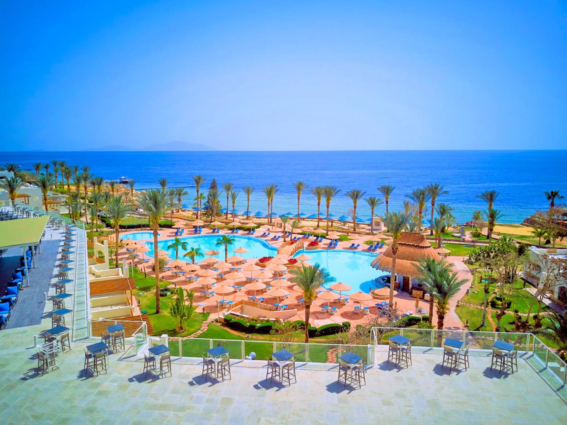فندق رويال جراند شرم ريزورت شرم الشيخ المظهر الخارجي الصورة The photo shows a beautiful resort scene with a spacious swimming pool at the center, surrounded by sun loungers and palm trees. There are areas for relaxation with tables and chairs positioned around the pool. In the background, the vibrant blue oce