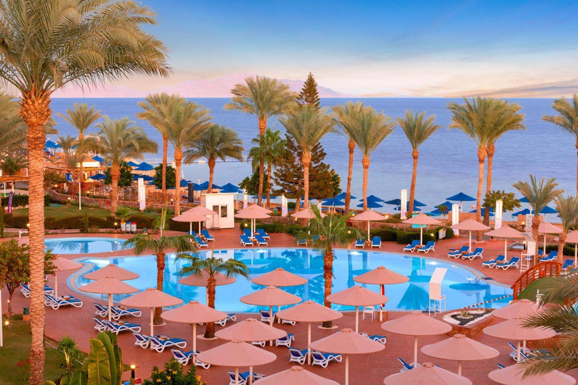 فندق رويال جراند شرم ريزورت شرم الشيخ المظهر الخارجي الصورة The photo shows a beautiful resort scene with a swimming pool surrounded by lounge chairs and umbrellas. Palm trees line the area, adding to the tropical atmosphere. In the background, there is a view of the sea under a clear sky, suggesting a peacef
