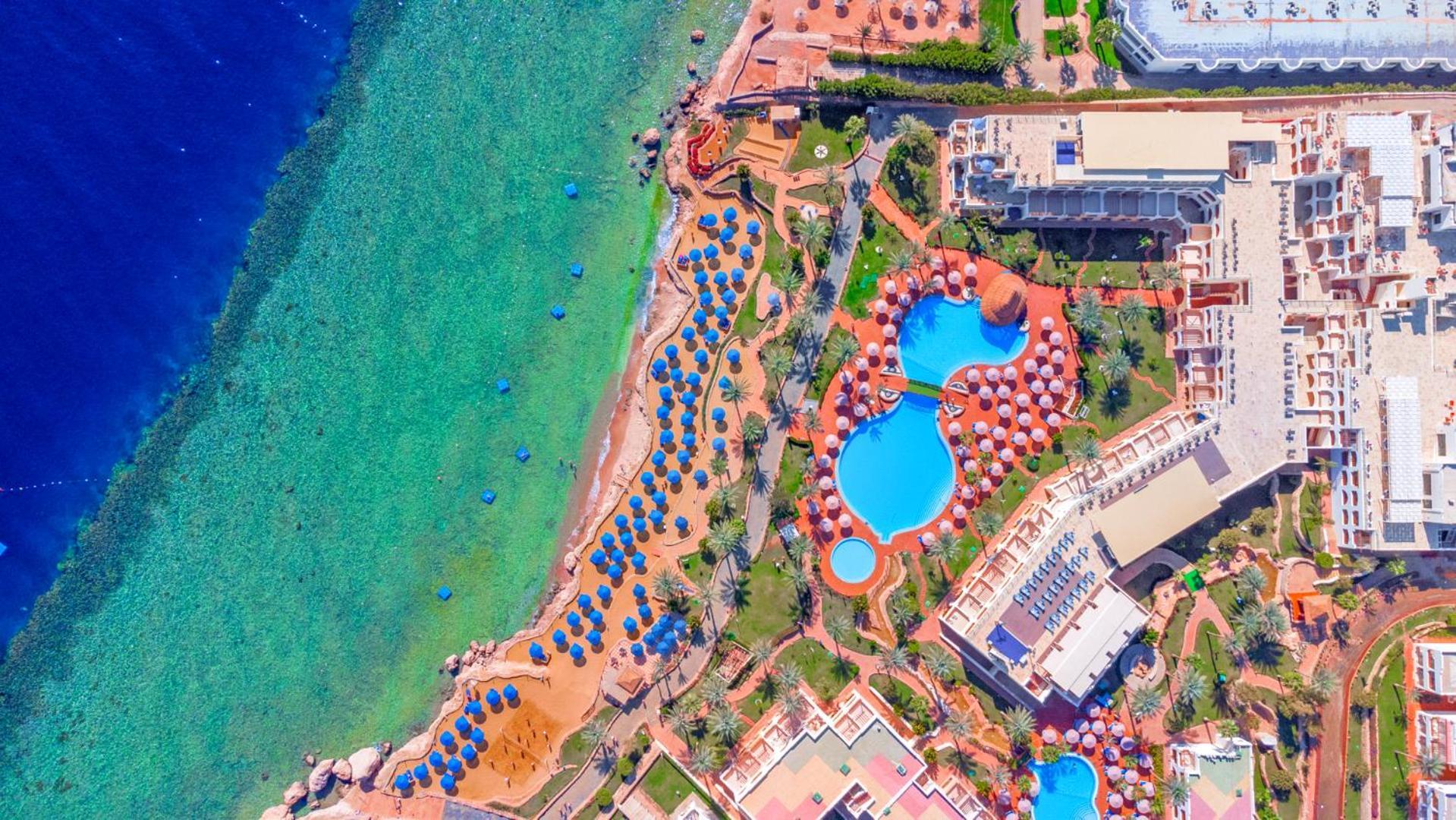 فندق رويال جراند شرم ريزورت شرم الشيخ المظهر الخارجي الصورة The photo showcases an aerial view of a resort area by the sea. It features a turquoise body of water alongside a sandy beach. There are several umbrellas set up on the beach and lounging areas. A large pool is visible, characterized by its unique sh