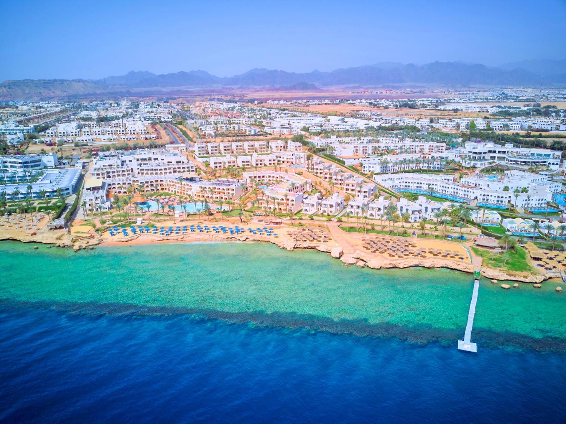 فندق رويال جراند شرم ريزورت شرم الشيخ المظهر الخارجي الصورة The photo shows an aerial view of a coastal resort area. In the foreground, there is a clear blue ocean with a gradient of colors from deep blue to lighter shades near the shore. The beach features sandy areas lined with sun loungers and umbrellas. F