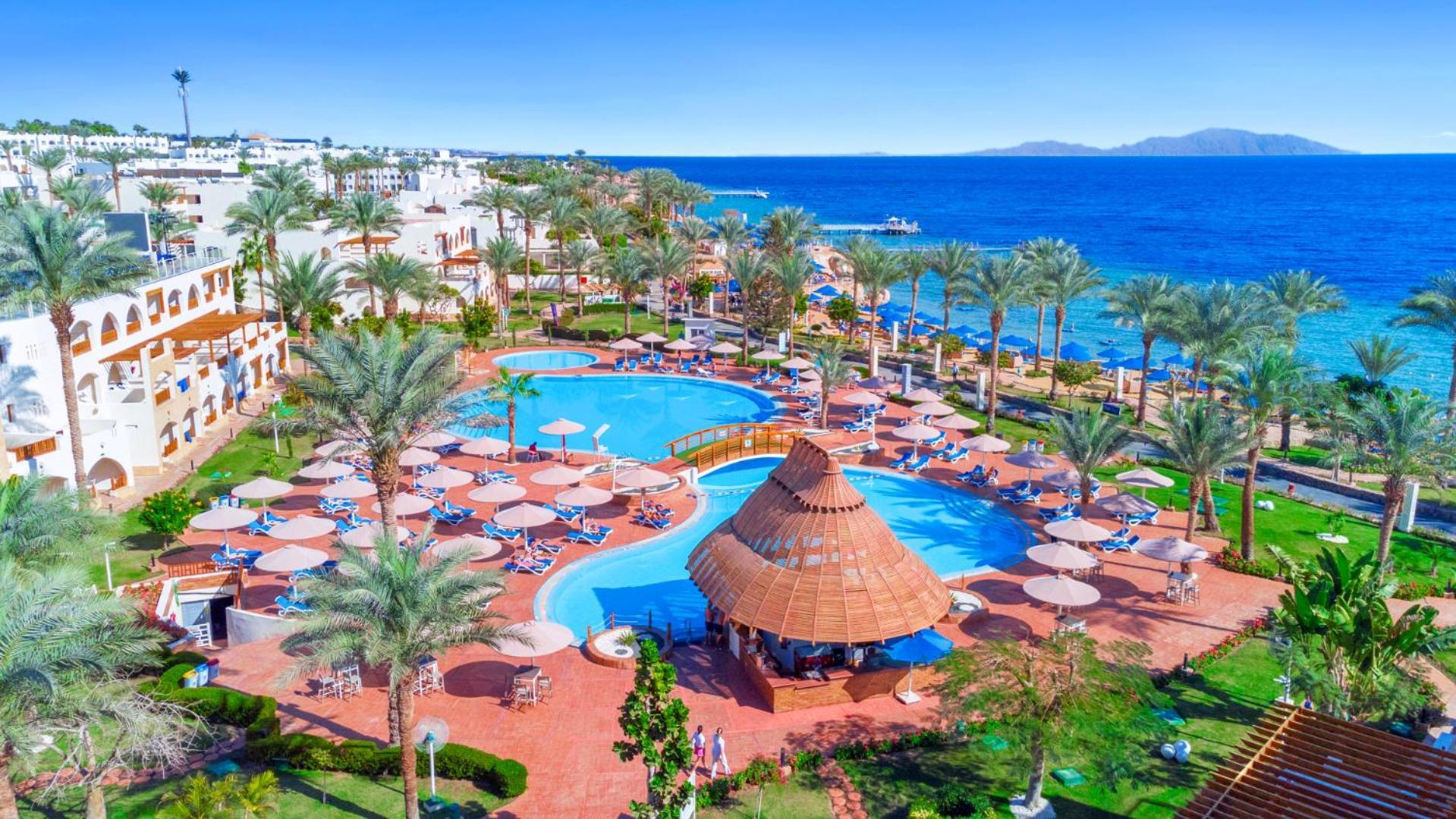 فندق رويال جراند شرم ريزورت شرم الشيخ المظهر الخارجي الصورة The image depicts a vibrant resort scene overlooking a clear blue sea. In the foreground, there are multiple swimming pools surrounded by sun loungers and umbrellas. A thatched-roof gazebo is situated near the center of the pool area. Palms and lush 