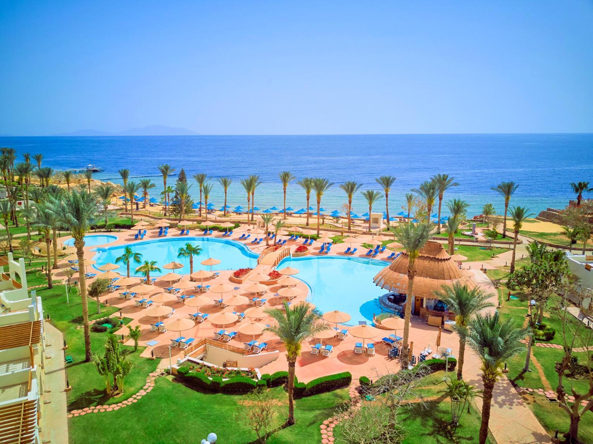 فندق رويال جراند شرم ريزورت شرم الشيخ المظهر الخارجي الصورة The photo depicts a beautiful resort scene by the ocean. In the foreground, there is a large swimming pool surrounded by palm trees and lounge chairs. The pool area features sunbeds and umbrellas, suggesting a relaxing atmosphere for guests. In the b