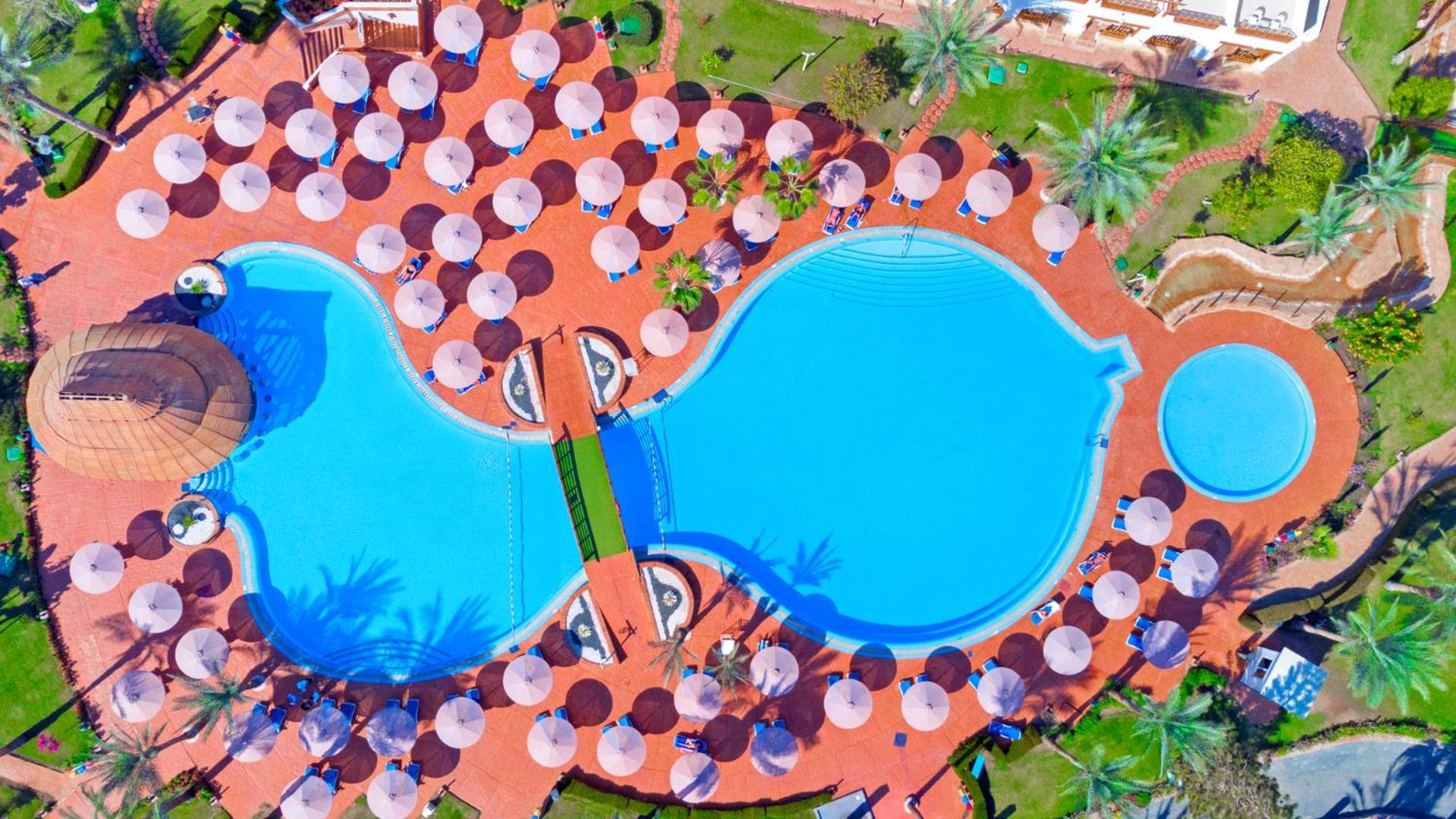 فندق رويال جراند شرم ريزورت شرم الشيخ المظهر الخارجي الصورة The photo shows an aerial view of a large swimming pool area, shaped in a distinctive layout resembling two interconnected pools. Surrounding the main pool are numerous umbrellas, providing shade for visitors. The pool area is set on a reddish-colore
