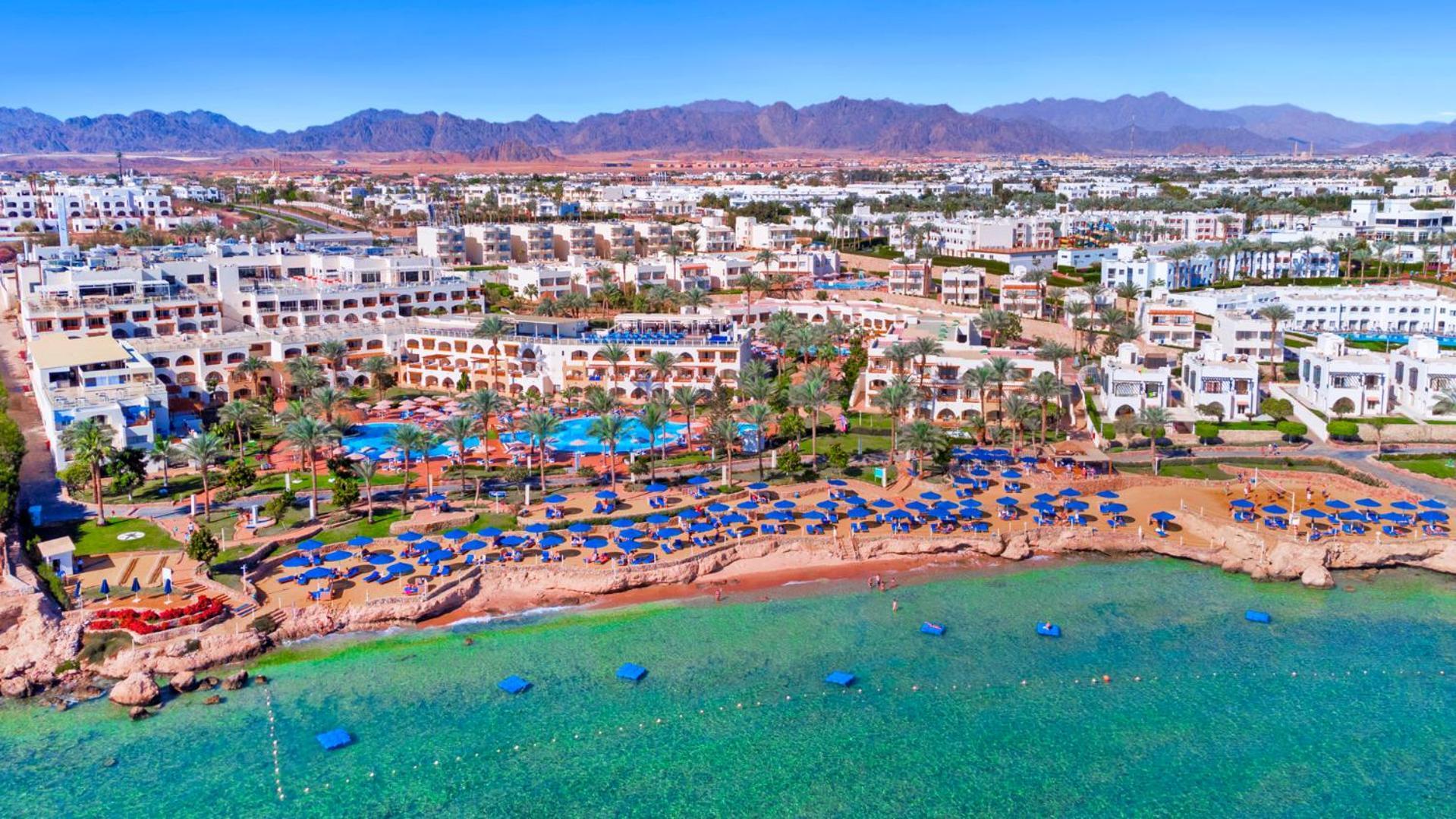 فندق رويال جراند شرم ريزورت شرم الشيخ المظهر الخارجي الصورة The photo shows an aerial view of a coastal resort area. In the foreground, there is a beach with greenish-blue water, where several blue beach umbrellas and lounges are set up for guests. The shoreline appears sandy, and there are some rocky areas. 