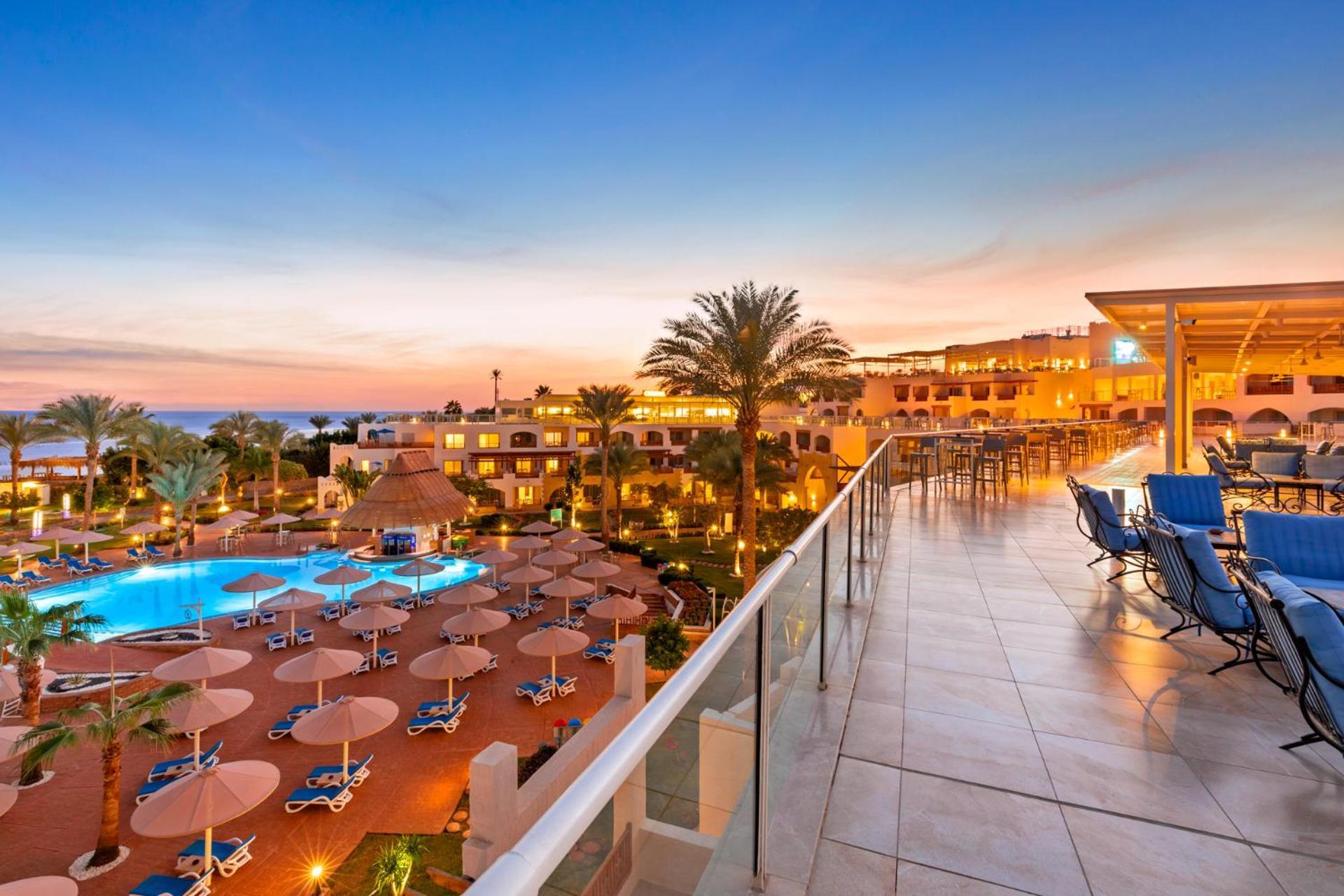 فندق رويال جراند شرم ريزورت شرم الشيخ المظهر الخارجي الصورة The photo depicts a scenic view of a resort during sunset. In the foreground, there is a balcony with tiled flooring, overlooking the resort. Below, a swimming pool is visible, surrounded by lounge chairs and large umbrellas, indicating a relaxing at