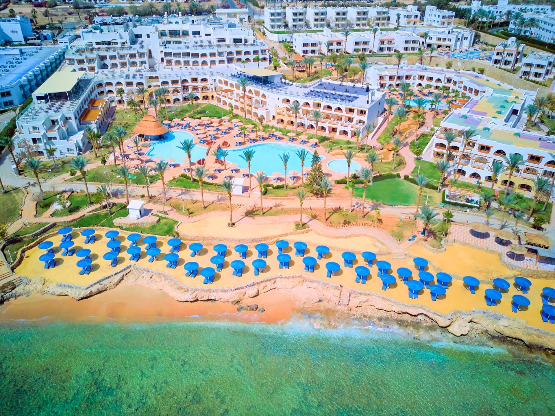 فندق رويال جراند شرم ريزورت شرم الشيخ المظهر الخارجي الصورة The photo shows a large resort complex situated near a beach. There is a turquoise swimming pool at the center, surrounded by green lawns and palm trees. Sun loungers with blue umbrellas are lined up on the sandy beach, and the beach area is adjacent