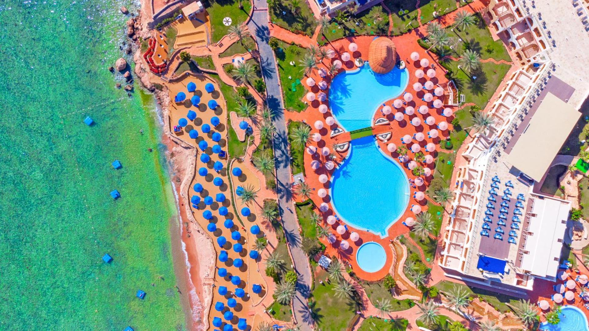 فندق رويال جراند شرم ريزورت شرم الشيخ المظهر الخارجي الصورة The photo shows an aerial view of a beach resort. In the foreground, there's a large swimming pool shaped like a flowing river, surrounded by lounge chairs and umbrellas. The pool has a distinctive, organic design with curved edges. To the left, ther