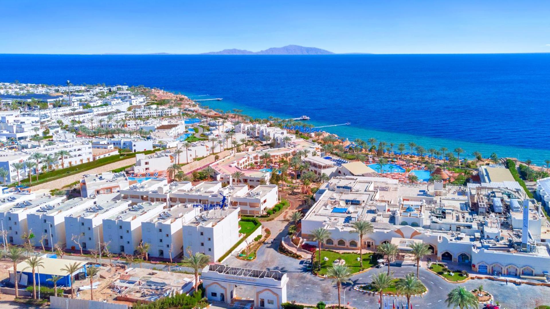 فندق رويال جراند شرم ريزورت شرم الشيخ المظهر الخارجي الصورة The photo shows a beautiful coastal landscape, featuring a resort area by the sea. There are white buildings, likely hotels or villas, arranged in a sprawling layout near the beach. The water is a vibrant blue, contrasting with the sandy shore and gr