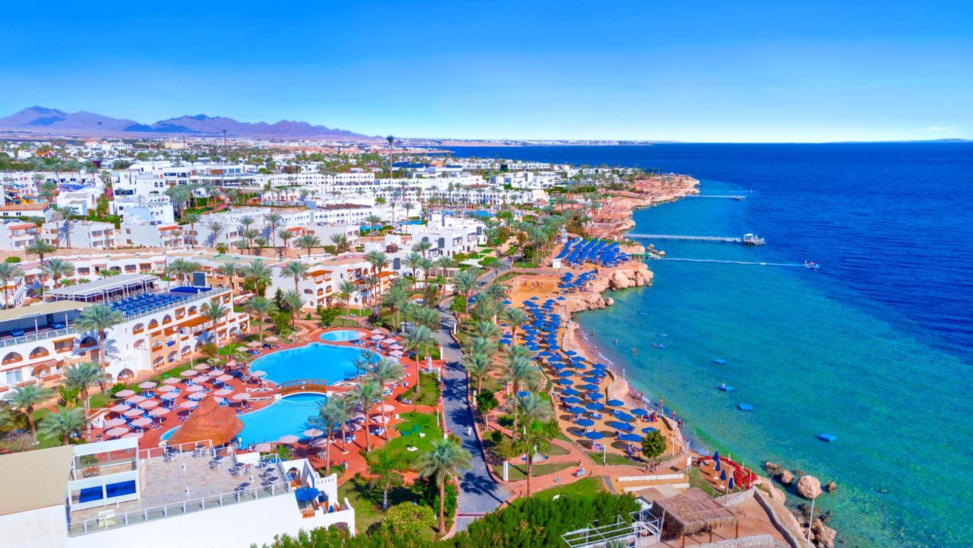 فندق رويال جراند شرم ريزورت شرم الشيخ المظهر الخارجي الصورة The photo features a picturesque coastal scene, likely from a resort area. In the foreground, there is a large swimming pool surrounded by lounge chairs. Further back, the landscape includes several buildings, possibly hotels or apartments, that over