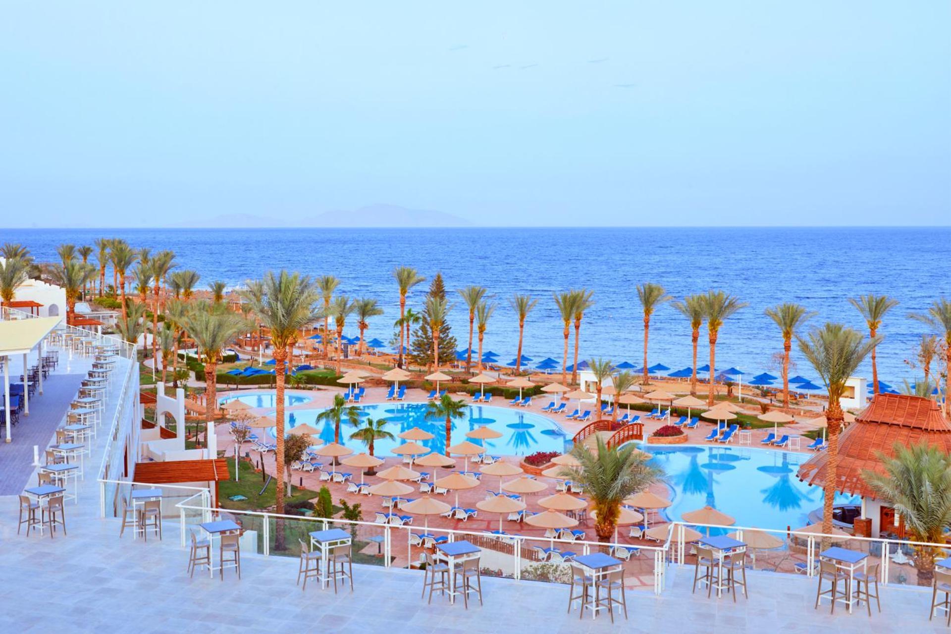 فندق رويال جراند شرم ريزورت شرم الشيخ المظهر الخارجي الصورة The photo depicts a scenic resort area by the sea. In the foreground, there is a large swimming pool surrounded by lounge chairs and umbrellas. Palm trees line the sides of the pool, adding to the tropical atmosphere. In the background, a beautiful b