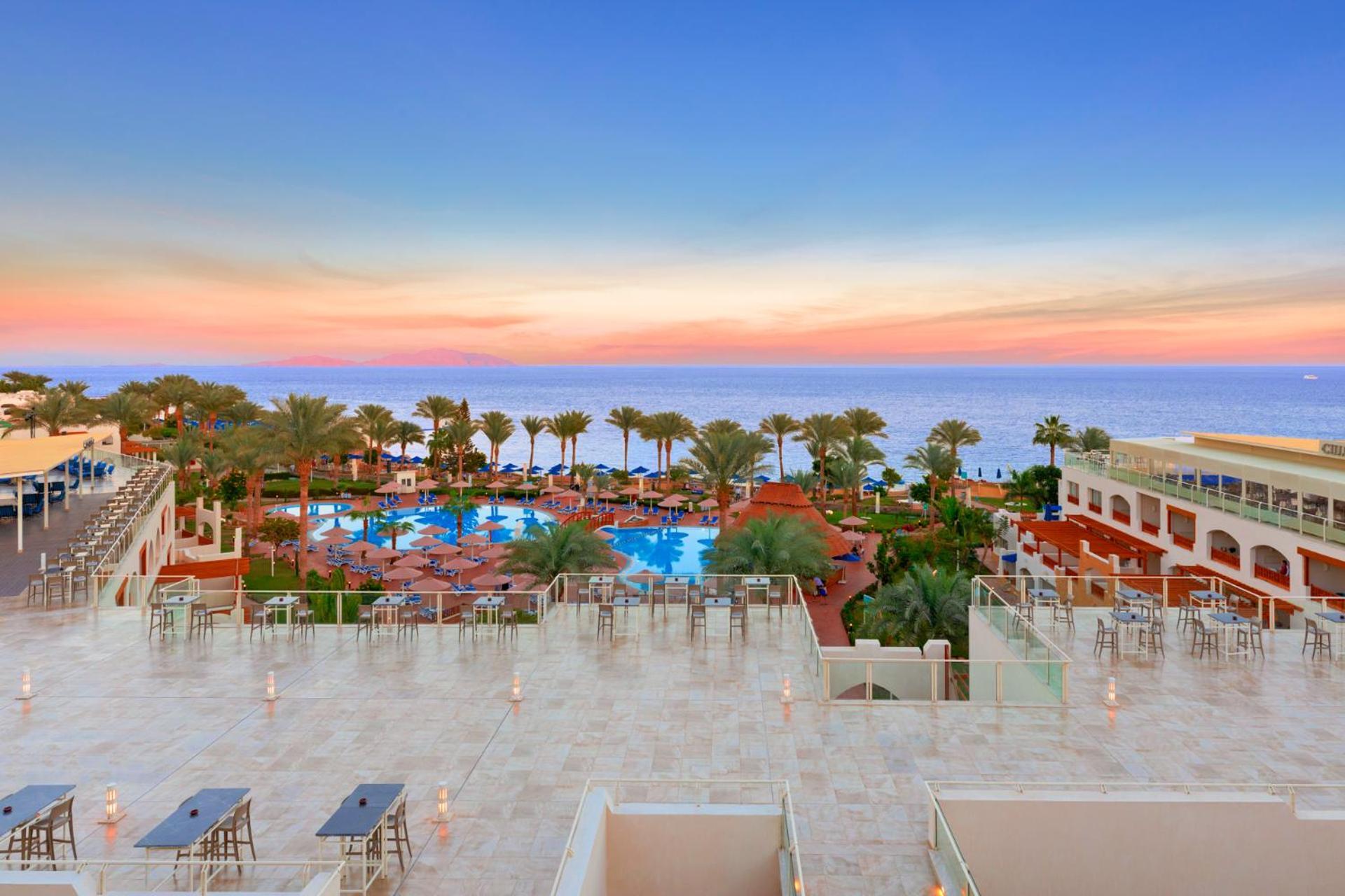 فندق رويال جراند شرم ريزورت شرم الشيخ المظهر الخارجي الصورة The photo depicts a beautiful resort scene at sunset. In the foreground, there is a spacious terrace area with modern furniture. Below, a large swimming pool is surrounded by lush palm trees and lounge chairs, indicating a relaxing atmosphere. The vi