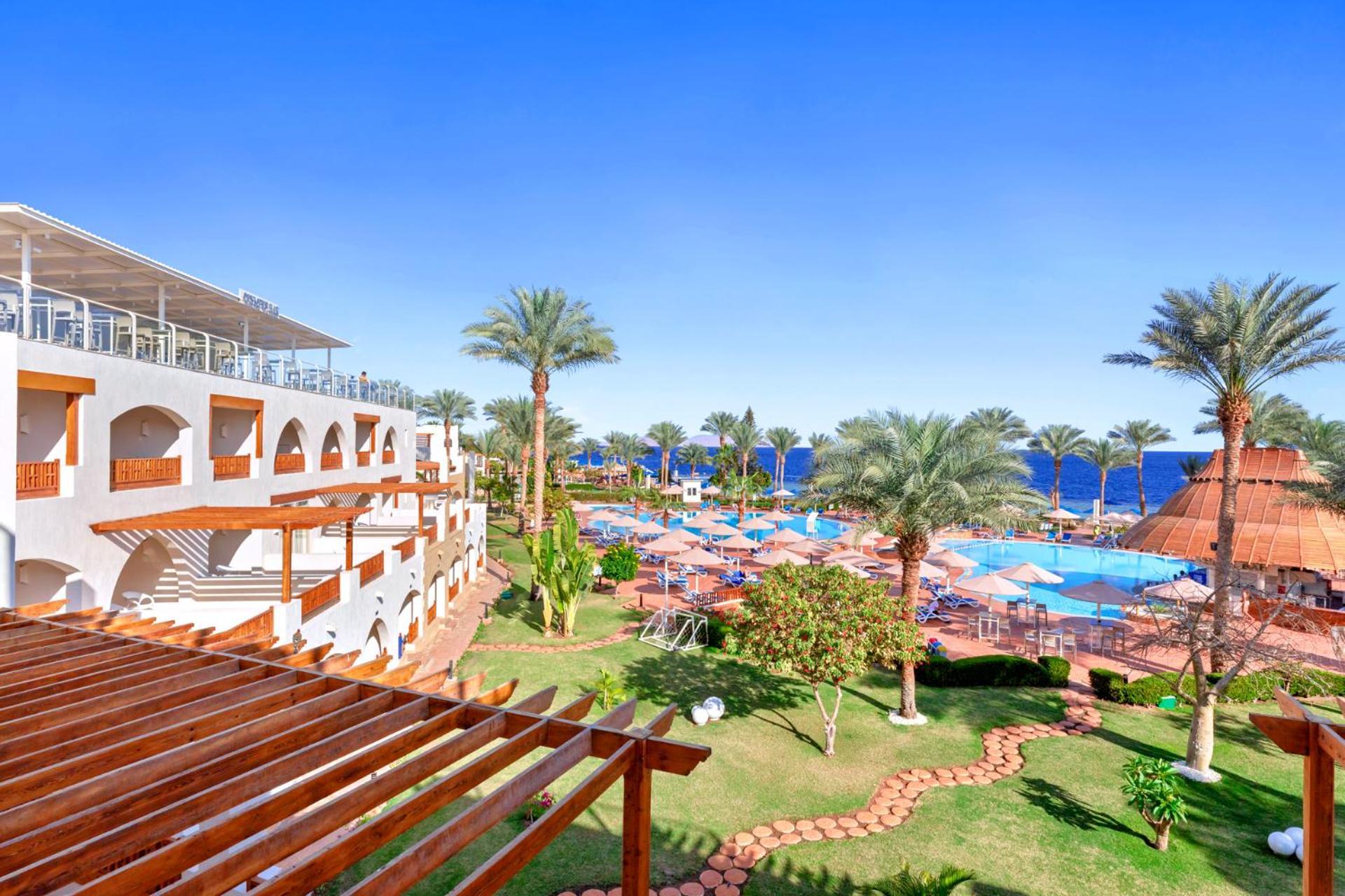 فندق رويال جراند شرم ريزورت شرم الشيخ المظهر الخارجي الصورة The photo showcases a beautiful resort setting, featuring several palm trees and a clear blue sky. In the foreground, there's an area with landscaped gardens, including green grass and a stone pathway. The resort buildings, with archways and wooden d