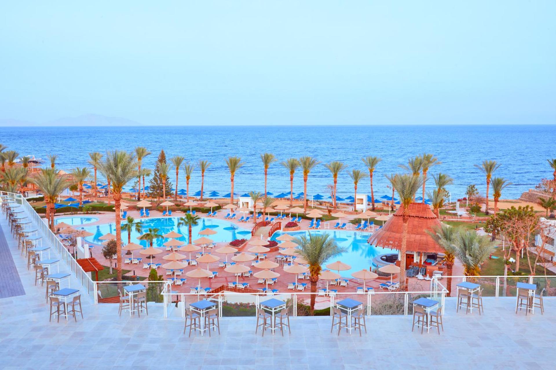 فندق رويال جراند شرم ريزورت شرم الشيخ المظهر الخارجي الصورة The photo shows a scenic view of a resort or beach area. In the foreground, there are several seating areas with bar stools on a terrace. Below, a large swimming pool is visible, surrounded by lounging chairs and umbrellas. Palm trees line the area, 