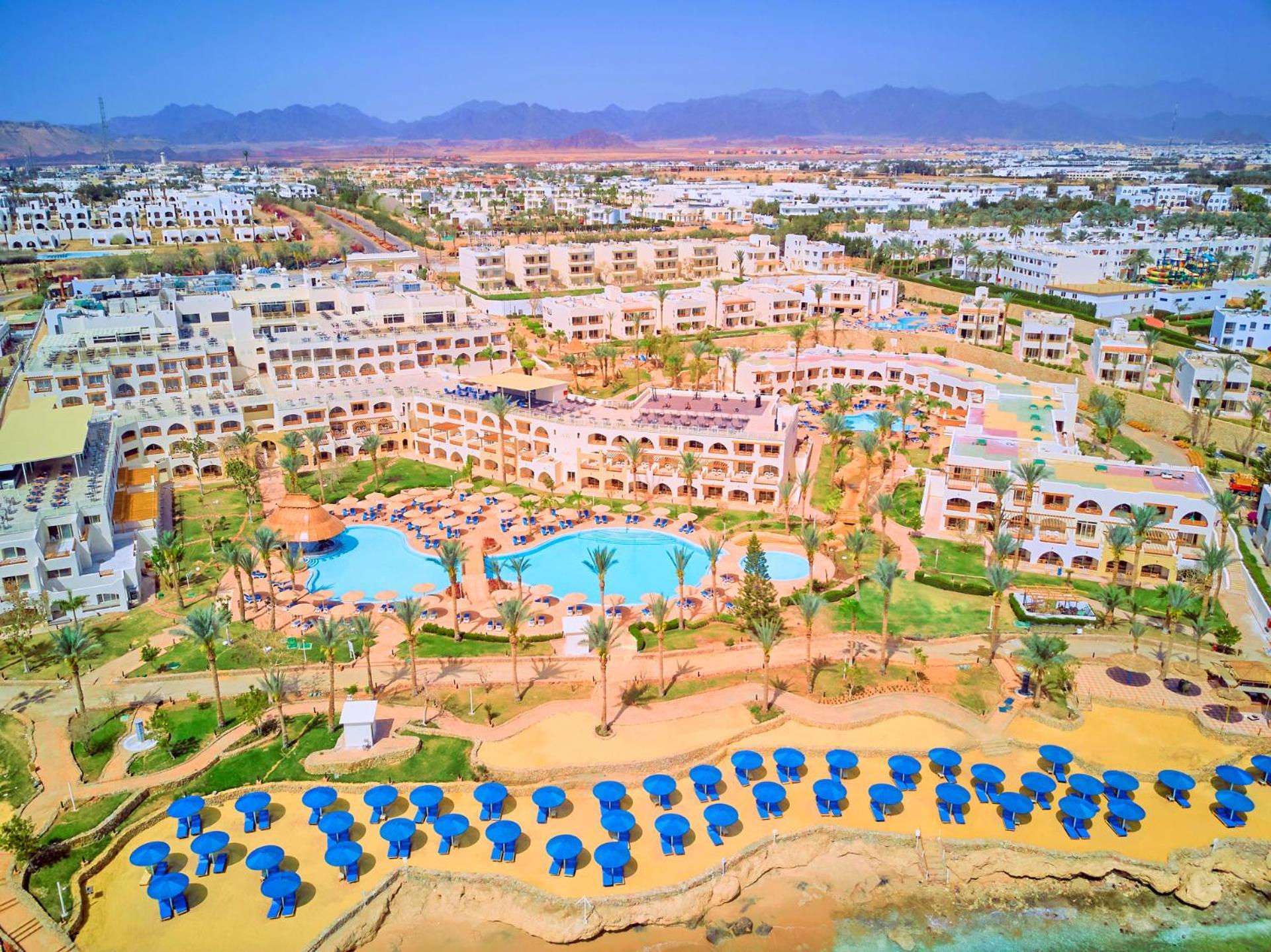 فندق رويال جراند شرم ريزورت شرم الشيخ المظهر الخارجي الصورة The photo shows a large resort complex set in a scenic location, likely near a beach. The layout features multiple buildings with a Mediterranean style of architecture, surrounded by lush greenery and palm trees. Central to the resort is a swimming p
