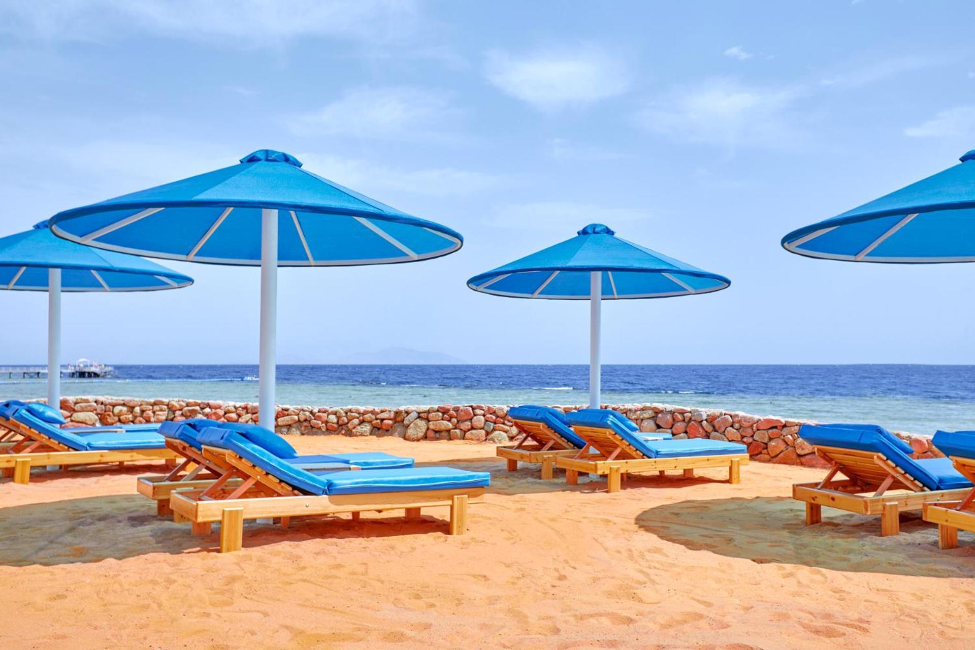 فندق رويال جراند شرم ريزورت شرم الشيخ المظهر الخارجي الصورة The photo shows a beach scene featuring several blue umbrellas providing shade over wooden lounge chairs. The sandy beach extends toward a calm sea, creating a relaxing atmosphere. The sky is mostly clear, with a few clouds, suggesting pleasant weath