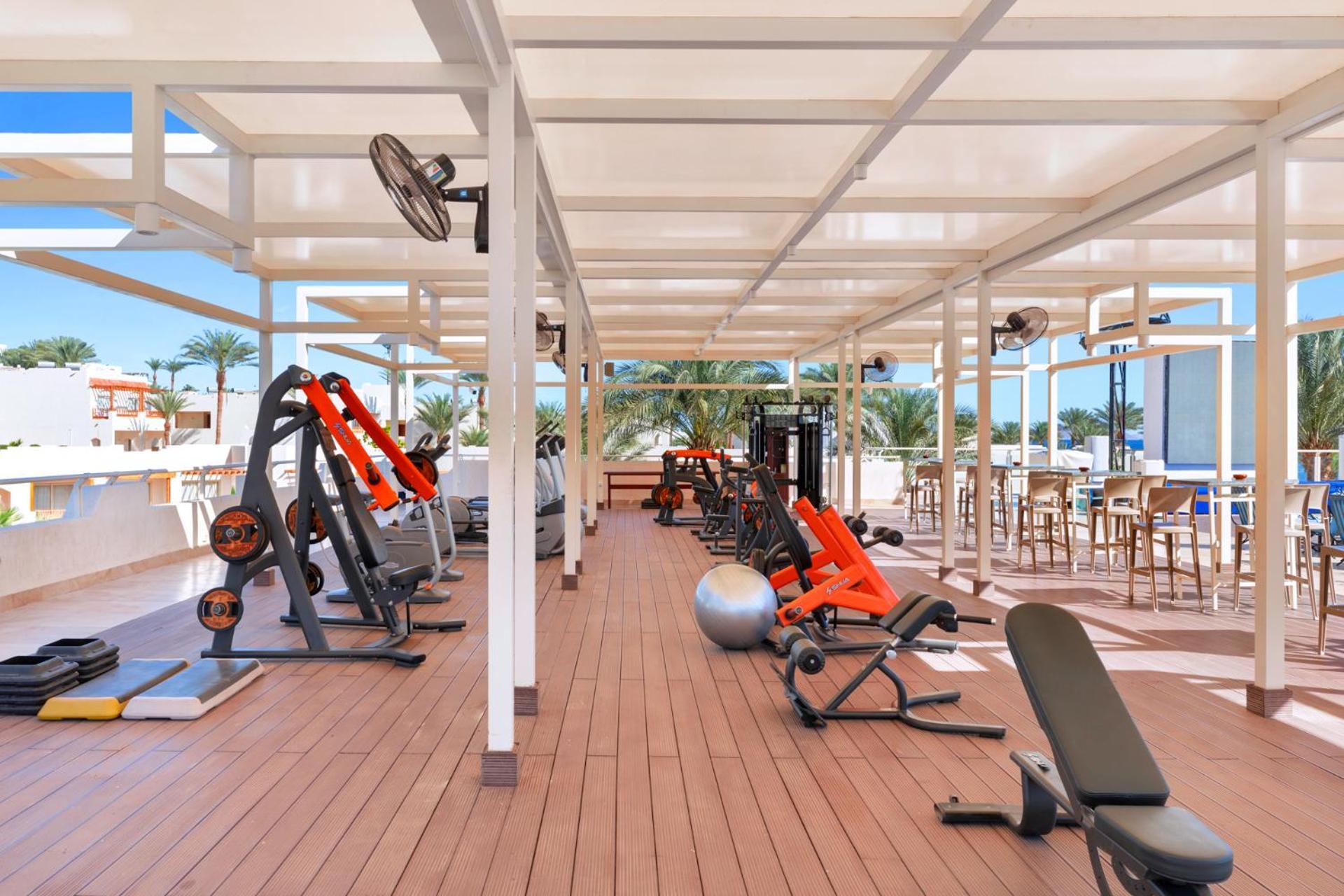 فندق رويال جراند شرم ريزورت شرم الشيخ المظهر الخارجي الصورة The photo depicts a gym area located outdoors or in a covered terrace, featuring various fitness equipment. There are several weight machines and benches, as well as some free weights. Large fans are installed for ventilation, and the space is design