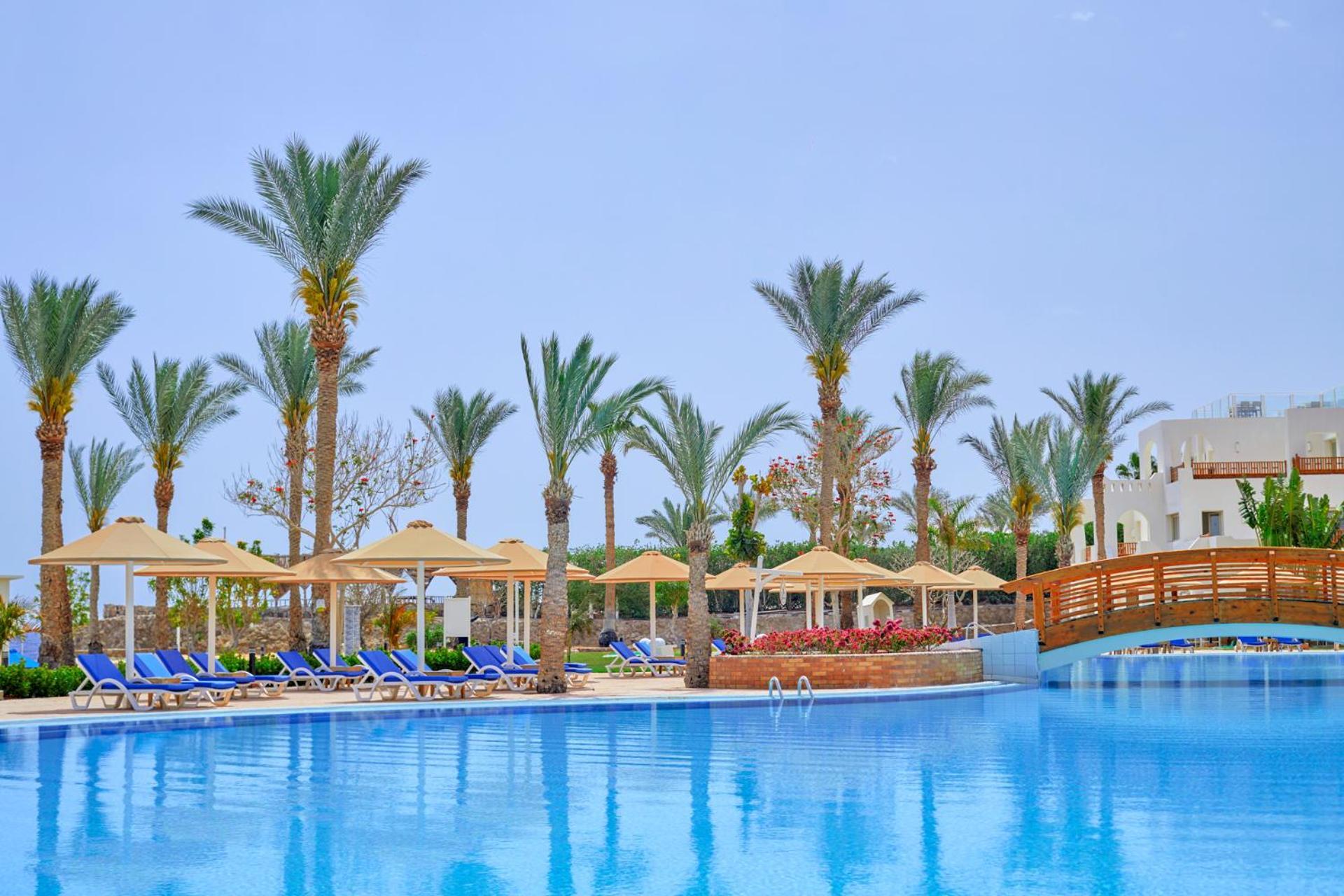 فندق رويال جراند شرم ريزورت شرم الشيخ المظهر الخارجي الصورة The photo shows a serene poolside scene at a resort. There is a large, clear blue pool reflecting the sky, surrounded by lounge chairs with blue cushions. Palm trees line the area, adding a tropical feel. Several covered loungers are placed under umb