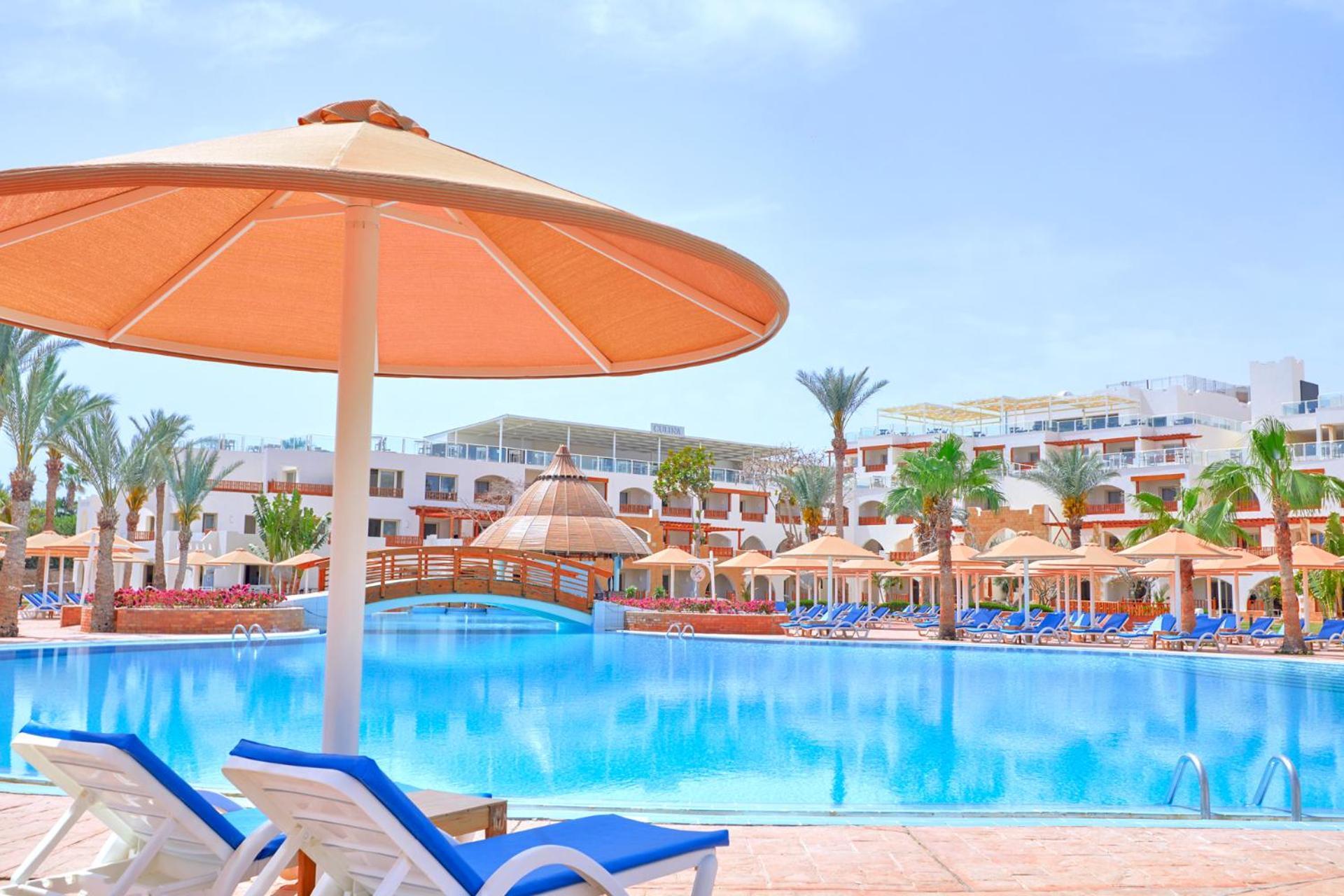 فندق رويال جراند شرم ريزورت شرم الشيخ المظهر الخارجي الصورة The photo shows a picturesque resort scene featuring a large swimming pool surrounded by lounge chairs and umbrellas. The pool is shimmering under a bright blue sky, and there are several palm trees in the background, adding to the tropical atmospher