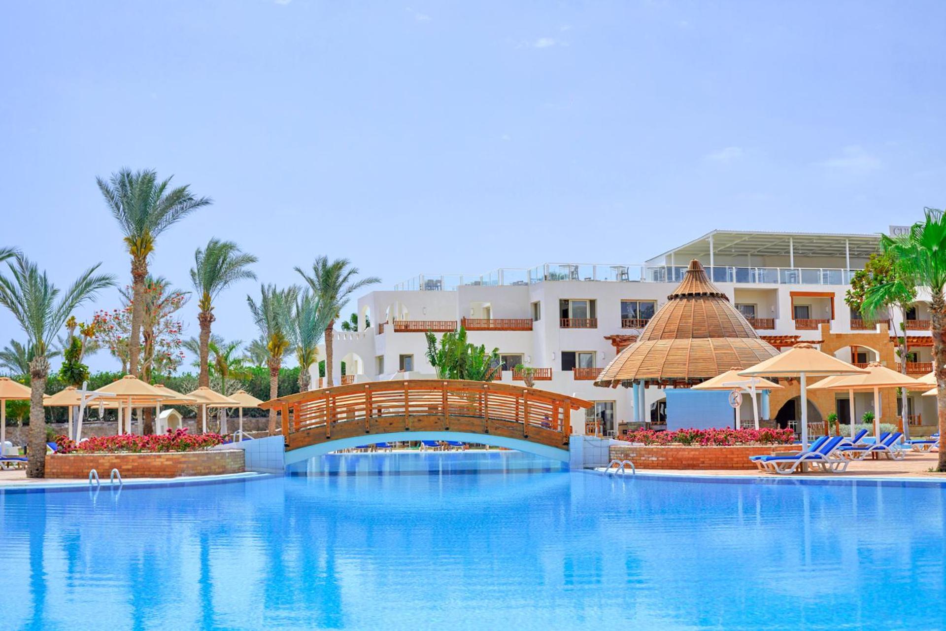 فندق رويال جراند شرم ريزورت شرم الشيخ المظهر الخارجي الصورة The photo depicts a serene resort setting featuring a large, calm swimming pool, which reflects the clear blue sky. In the foreground, a wooden bridge arches over the pool. Surrounding the pool are several palm trees and tropical foliage, contributin