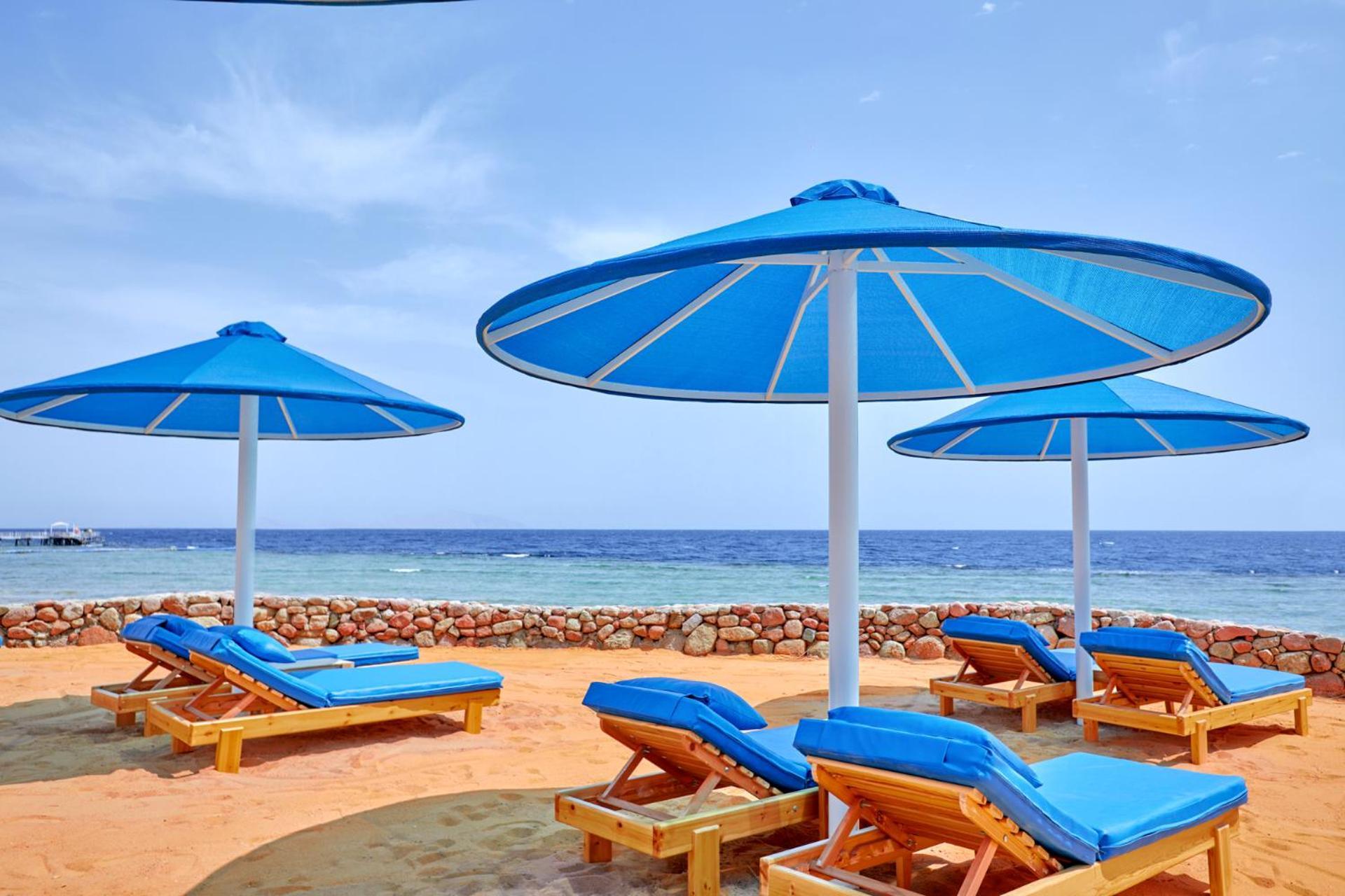 فندق رويال جراند شرم ريزورت شرم الشيخ المظهر الخارجي الصورة The photo shows a serene beach scene with several blue sun loungers arranged beneath large blue umbrellas. The loungers are positioned on sandy ground, with the ocean visible in the background. The sky is clear, indicating a bright and sunny day, and