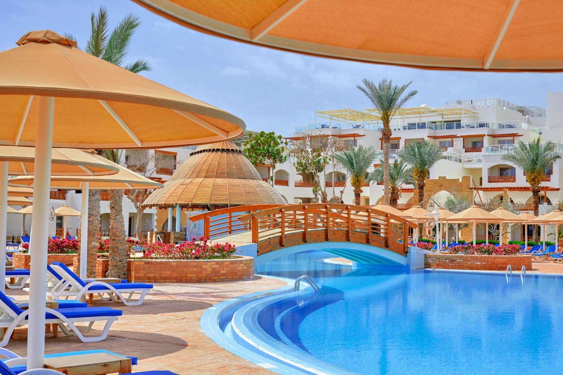 فندق رويال جراند شرم ريزورت شرم الشيخ المظهر الخارجي الصورة The photo shows a picturesque resort scene featuring a bright blue swimming pool surrounded by sun loungers and parasols. In the background, there is a wooden bridge arching over a section of the pool, leading towards the other side. The area is ador