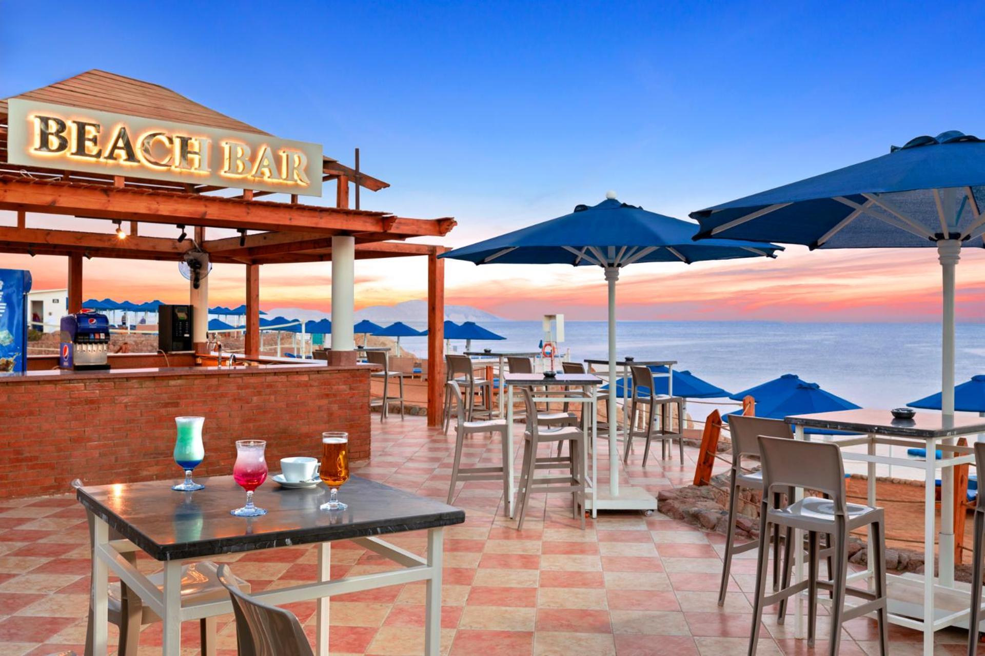 فندق رويال جراند شرم ريزورت شرم الشيخ المظهر الخارجي الصورة The photo shows a beach bar setting, featuring a wooden structure with the words "BEACH BAR" prominently displayed. There are several tables and chairs arranged on a tiled floor, with some drinks placed on the tables. Blue umbrellas provide shade for