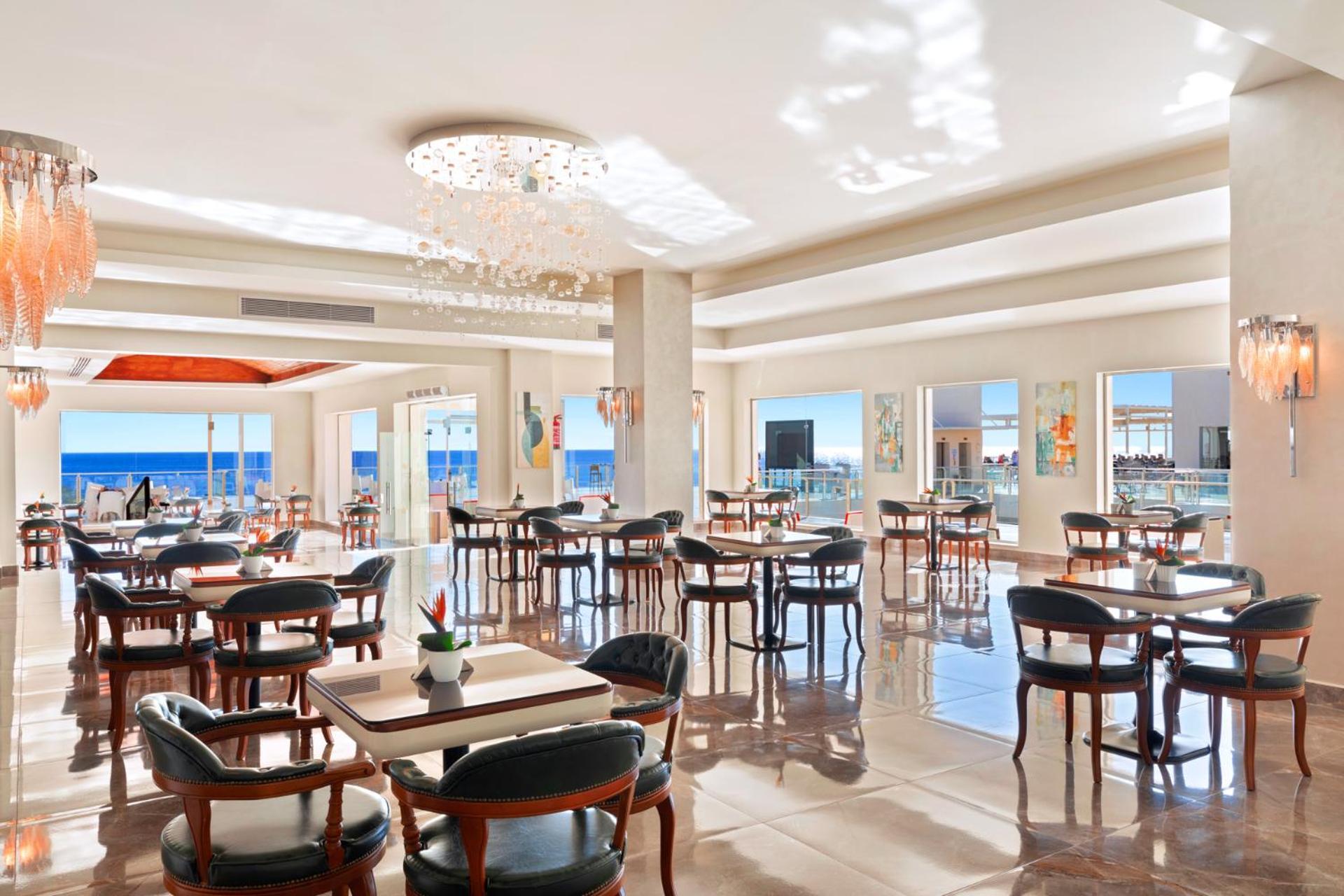 فندق رويال جراند شرم ريزورت شرم الشيخ المظهر الخارجي الصورة The photo shows a bright and elegant restaurant interior. There are several tables arranged throughout the space, each with chairs around them. The decor features modern lighting, including a chandelier, and large windows that allow natural light to 