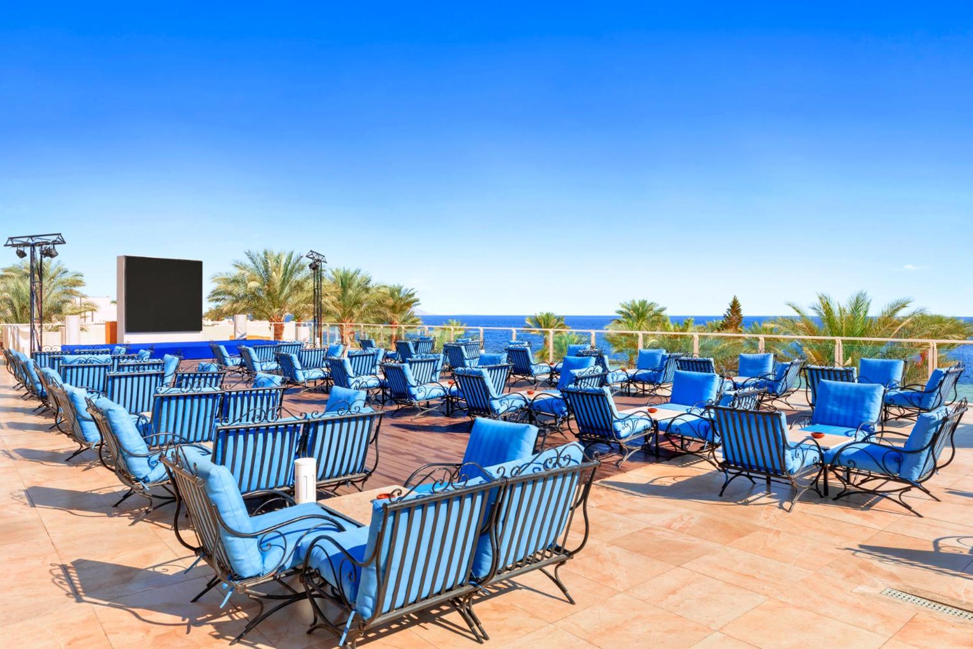 فندق رويال جراند شرم ريزورت شرم الشيخ المظهر الخارجي الصورة The photo features a sunny outdoor space with numerous blue lounge chairs arranged in rows on a tiled patio. In the background, there is a clear blue sky and a view of the ocean, along with some palm trees adding to the tropical atmosphere. Additiona