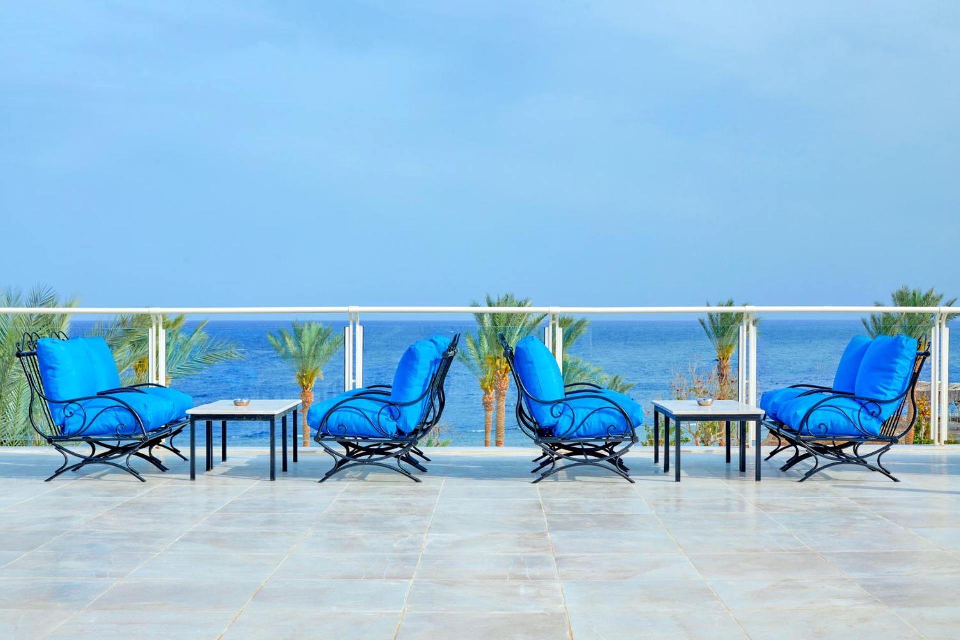 فندق رويال جراند شرم ريزورت شرم الشيخ المظهر الخارجي الصورة The photo depicts a serene outdoor patio area with four blue lounge chairs arranged around small tables. The setting features a clear view of the ocean with palm trees visible in the background, complementing the tranquil atmosphere. The overall colo