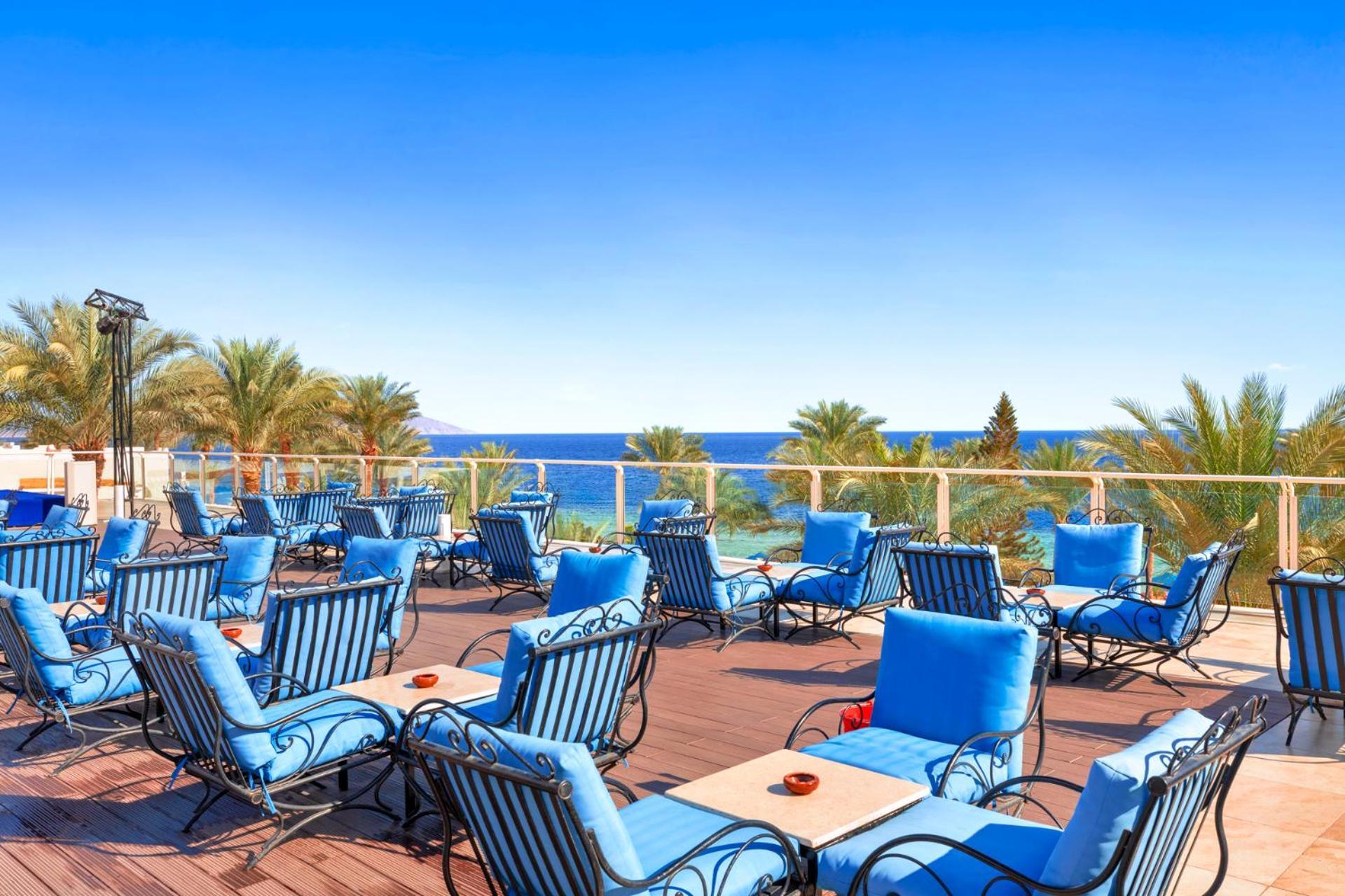 فندق رويال جراند شرم ريزورت شرم الشيخ المظهر الخارجي الصورة The photo shows an outdoor deck or terrace area, adorned with numerous blue lounge chairs arranged neatly. In the background, there are palm trees and a clear blue sea that stretches toward the horizon under a bright sky. The setting appears to be a 
